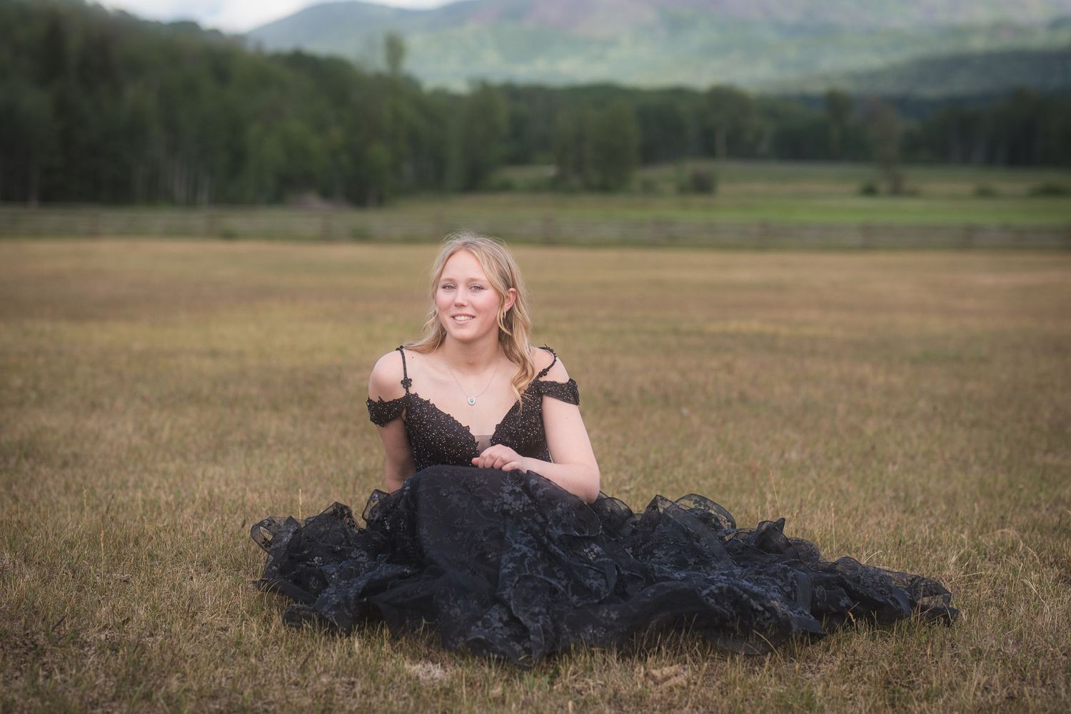 Blonde woman in black tulle gown sits gracefully in golden field with mountains behind.