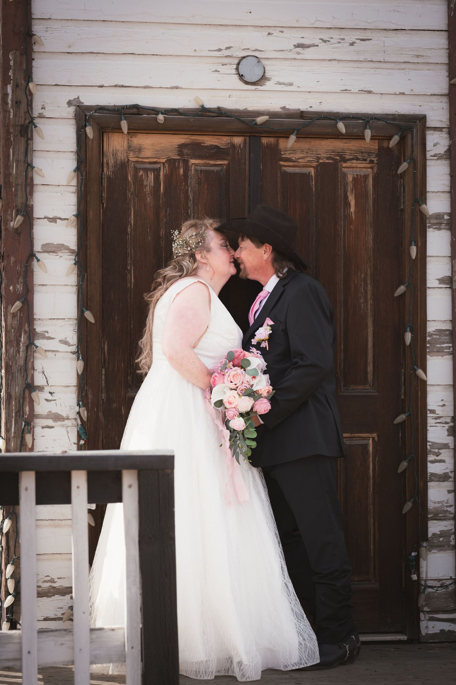 Newlywed couple shares first kiss in front of vintage wooden doors, bride holding pink rose bouquet.