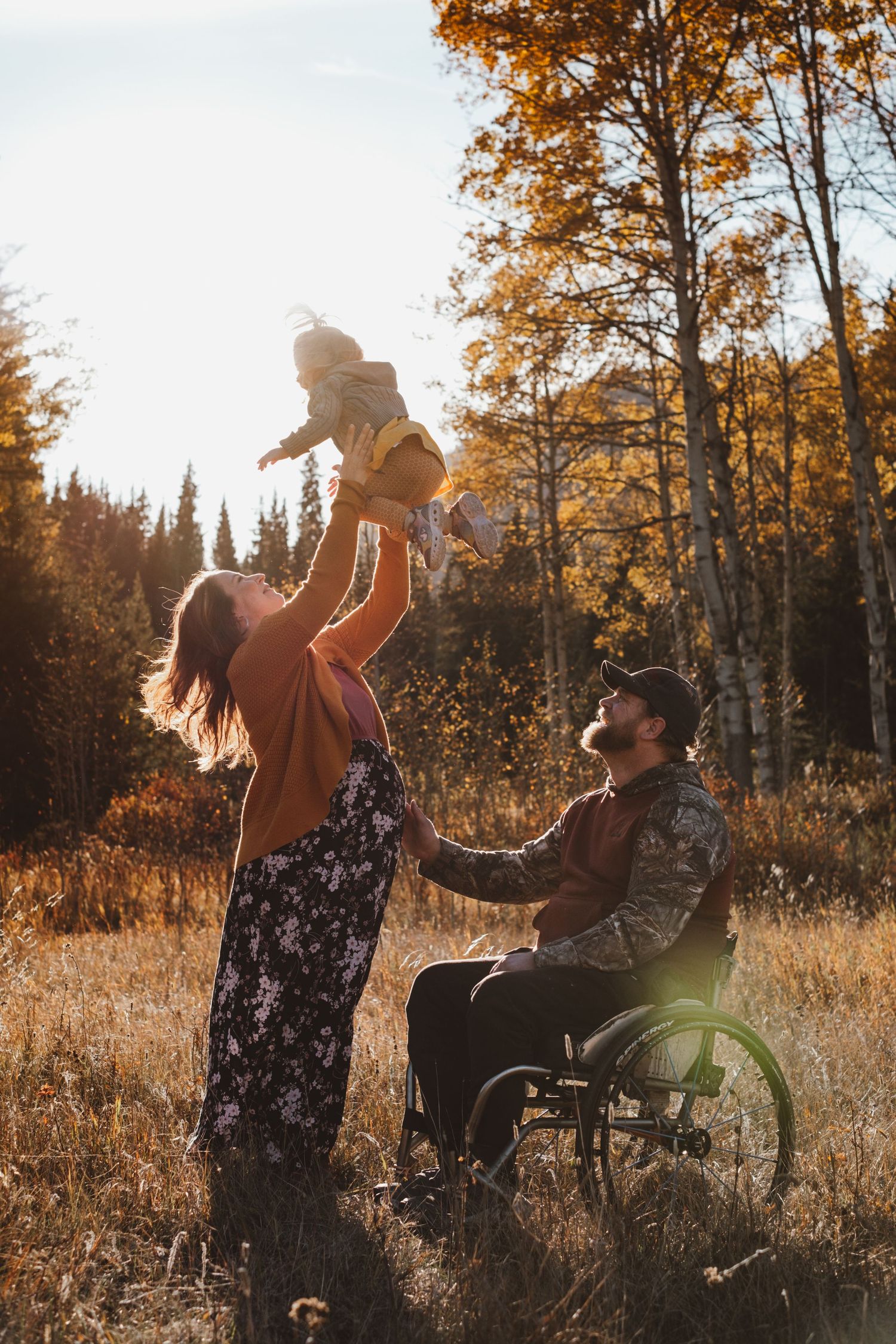 Mother lifting baby in the air beside father in wheelchair during a warm autumn family photo session outdoors.