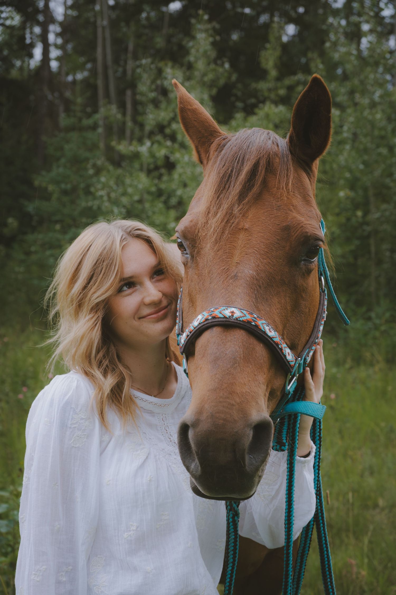 Smiling blonde woman in white blouse posing with a chestnut horse wearing a teal bridle in a green forest.