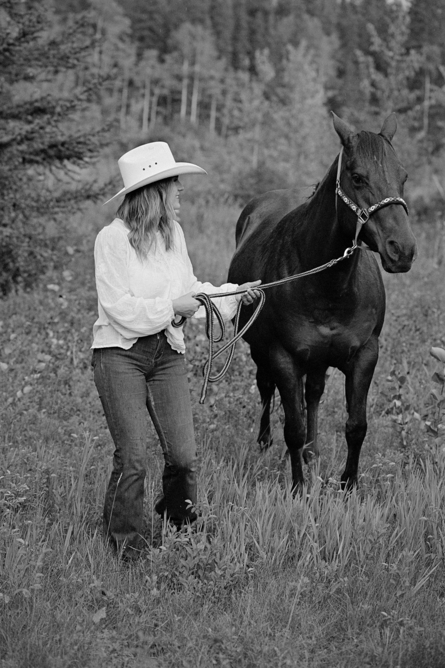 Woman in cowboy hat leading a dark horse through a grassy field in black and white photography.