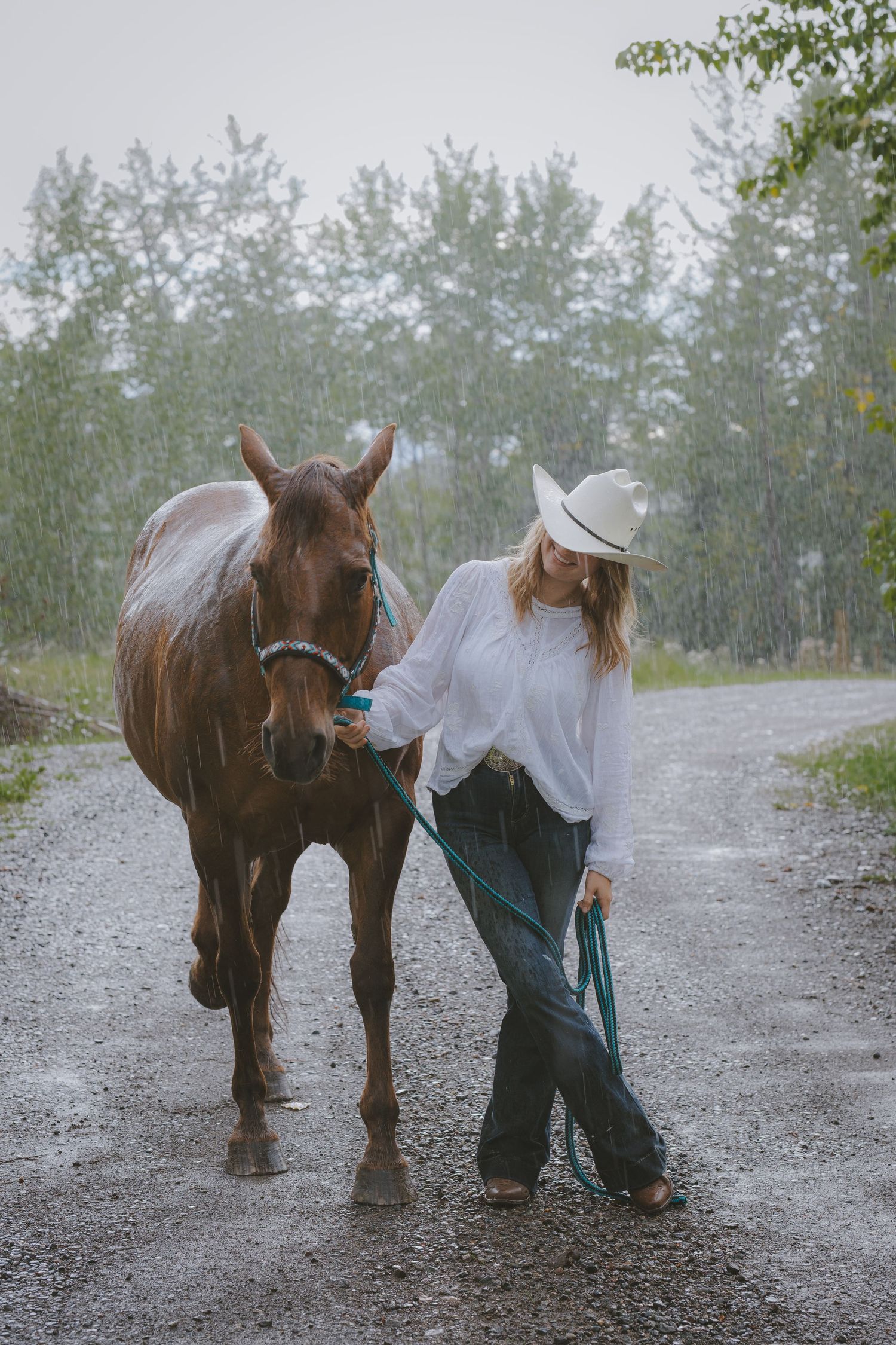 Woman in white shirt and cowboy hat leading a chestnut horse on a gravel path in the rain.