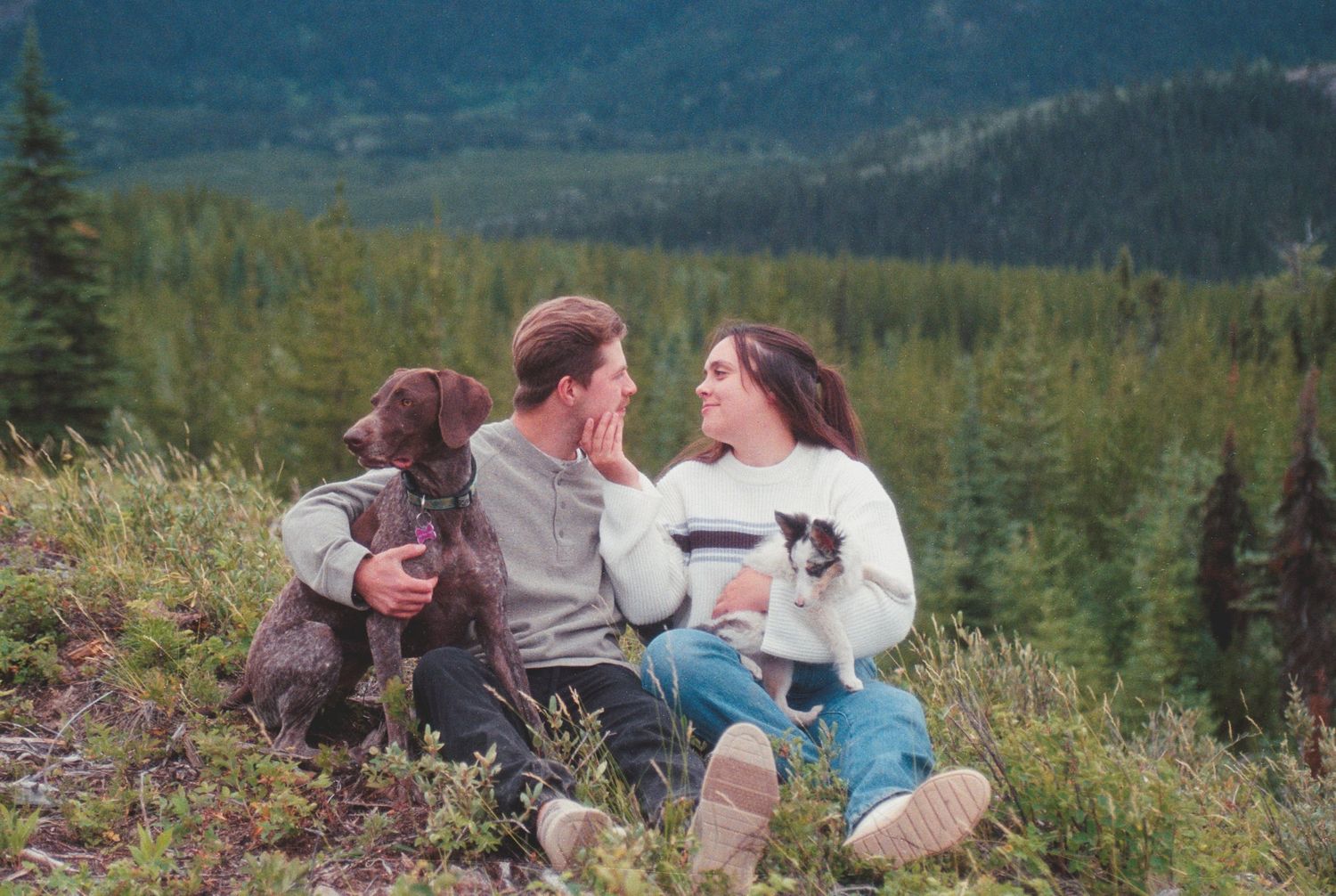 Couple sitting on a hillside with their dog, smiling at each other amid mountain scenery.