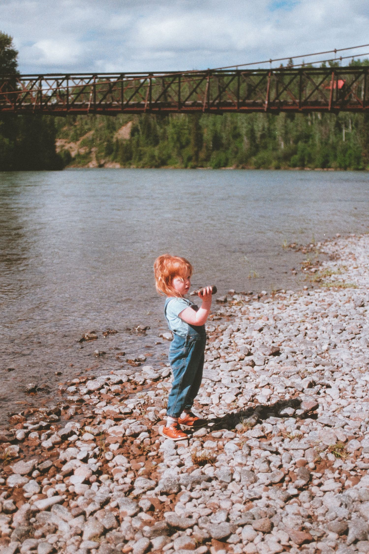 A young red-haired toddler in denim overalls stands on a rocky riverbank near an old iron bridge.