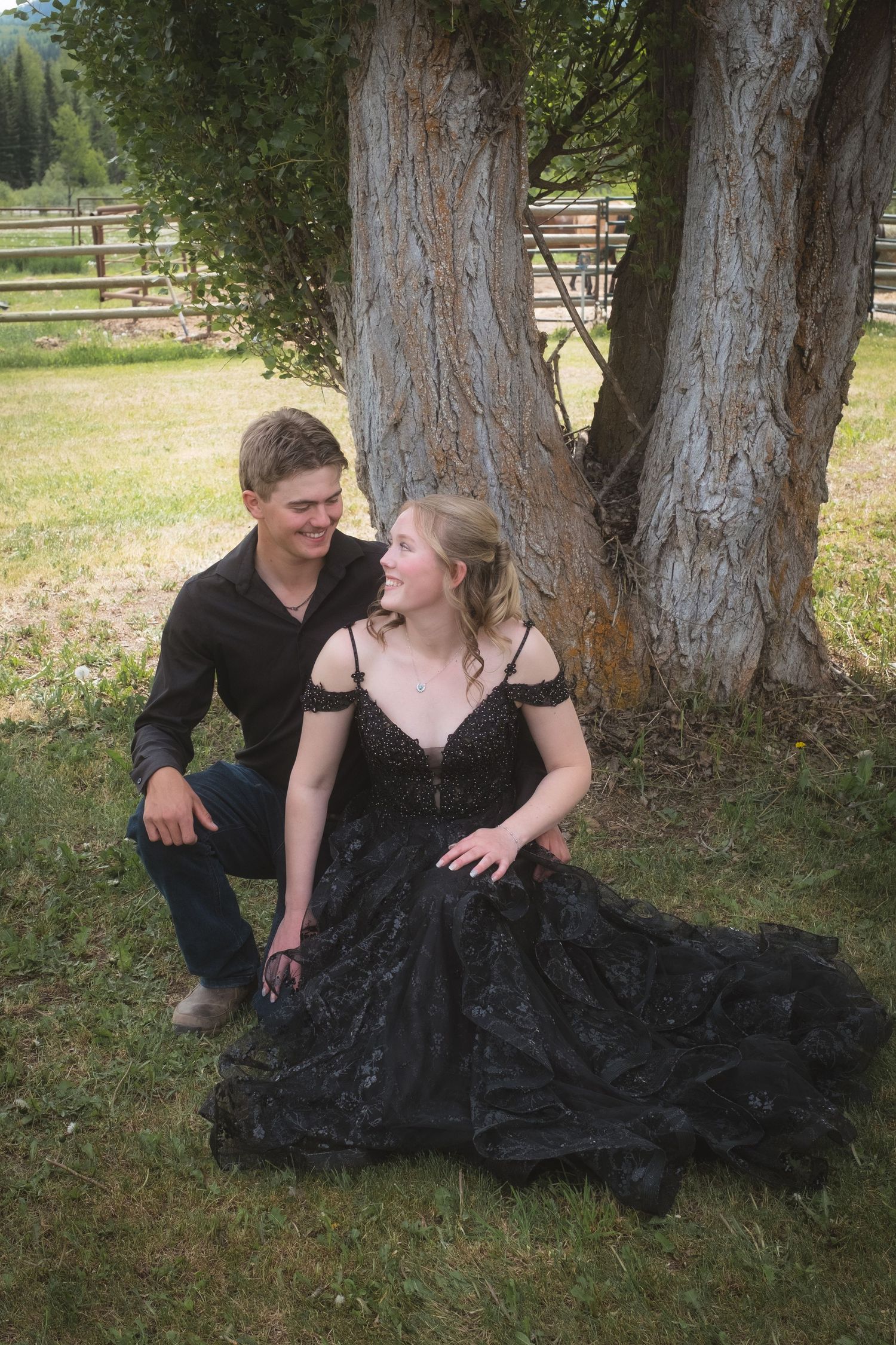 Young couple in formal attire sitting together under large tree on green grass at a ranch.