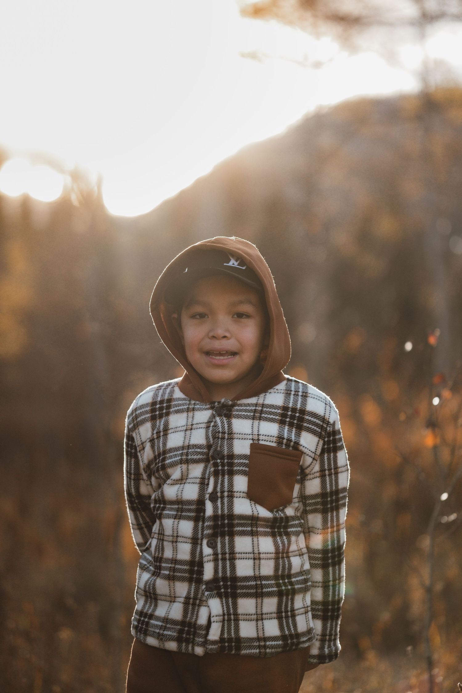 A young boy in a hooded plaid shirt smiles in a sunlit autumn outdoor setting.