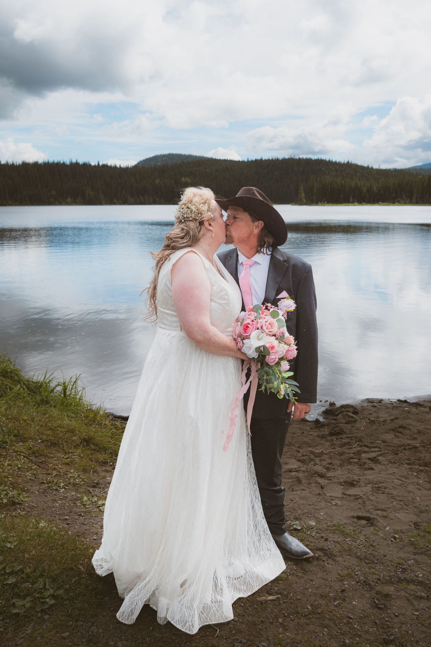 Bride and groom kissing by a lake with forested hills in background.