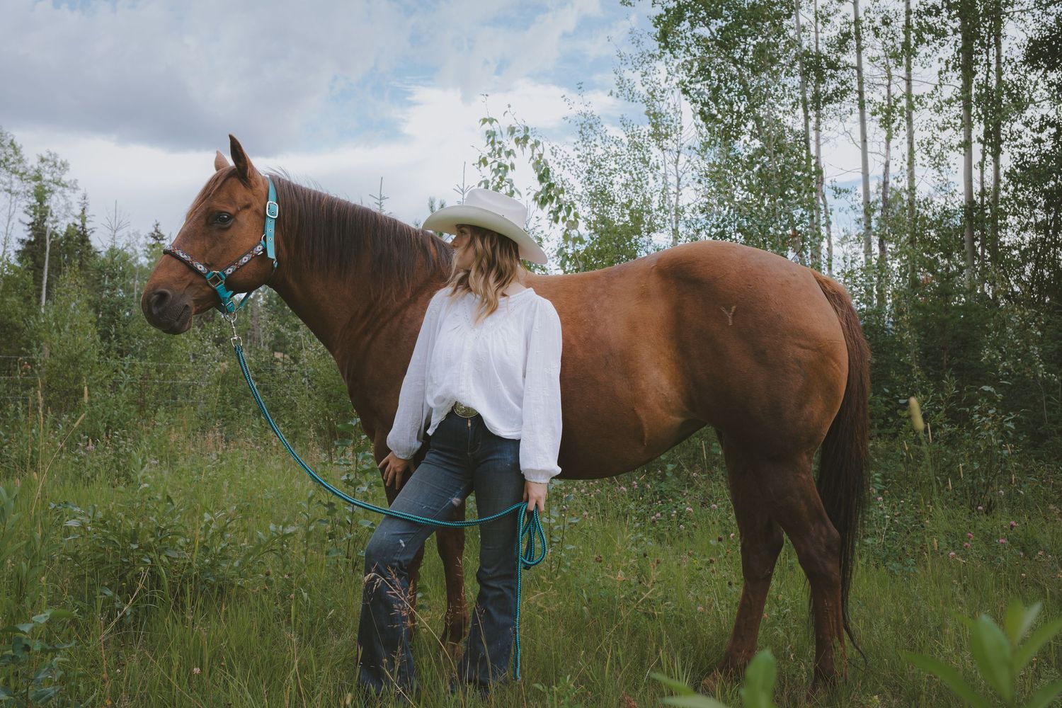 A woman in a white blouse and cowboy hat stands beside a chestnut horse in a lush green field.