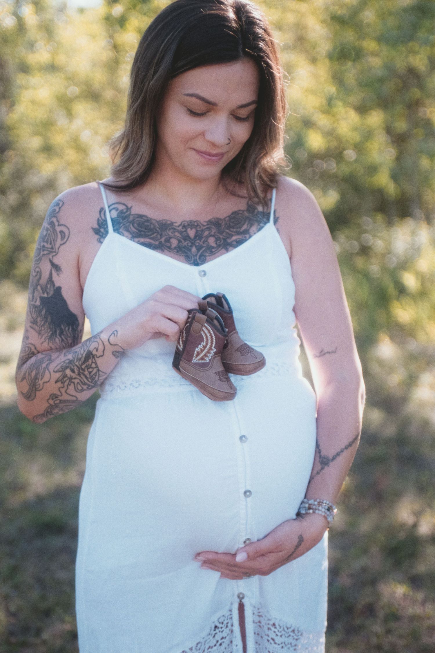 Tattooed pregnant woman in white dress holding tiny baby boots over her bump in a natural setting.