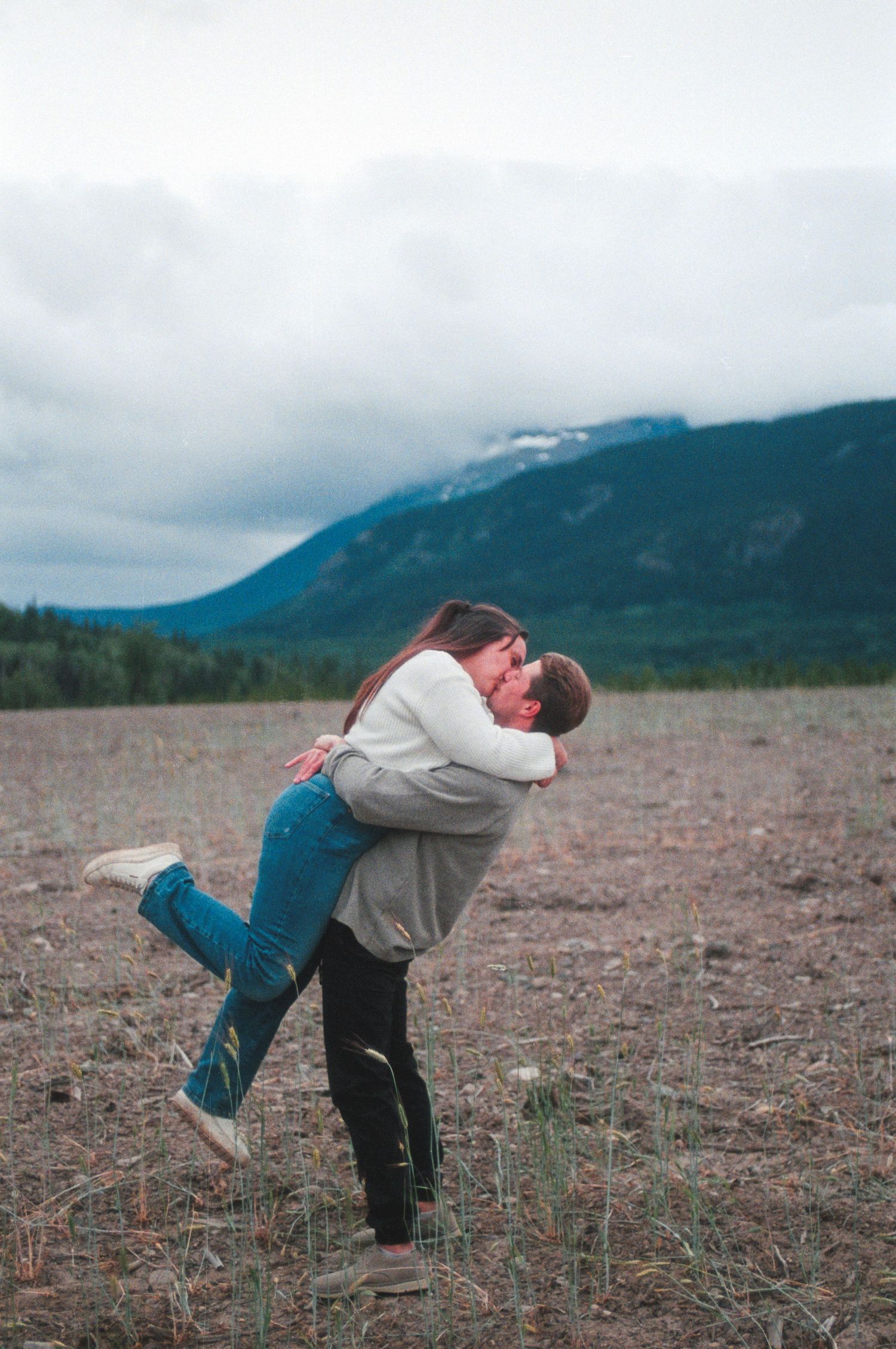 Couple sharing a romantic kiss in an open field with misty mountain backdrop on a cloudy day.