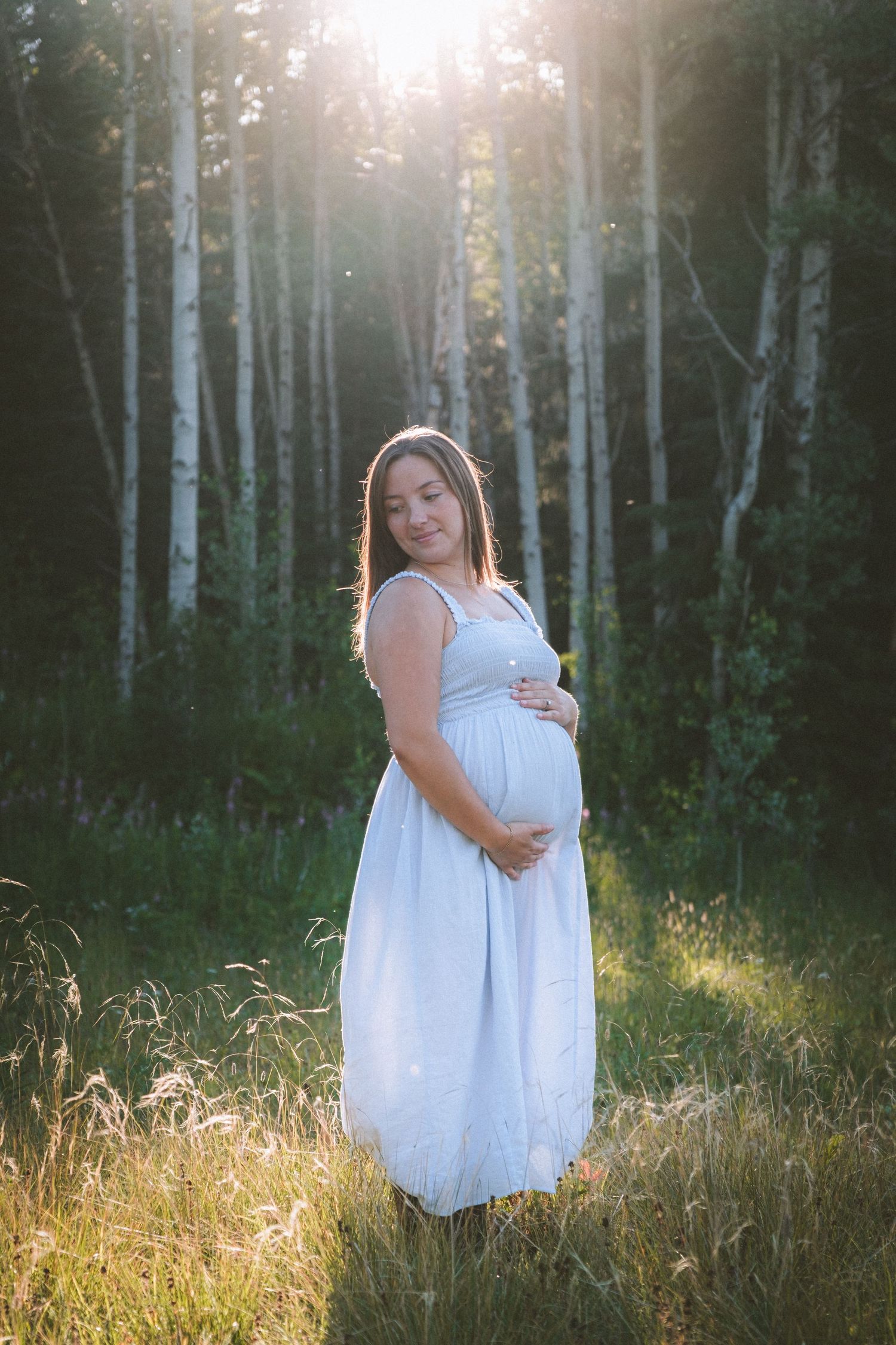 Pregnant woman in white dress cradling baby bump in sunlit forest clearing with birch trees.