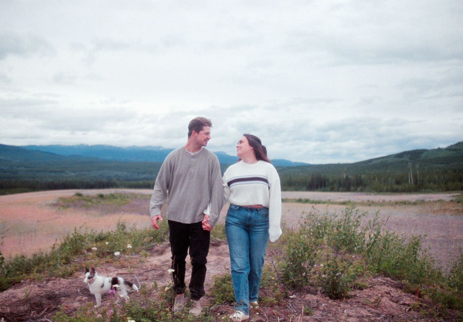 Happy couple walking hand in hand with their dog through a scenic mountain landscape.