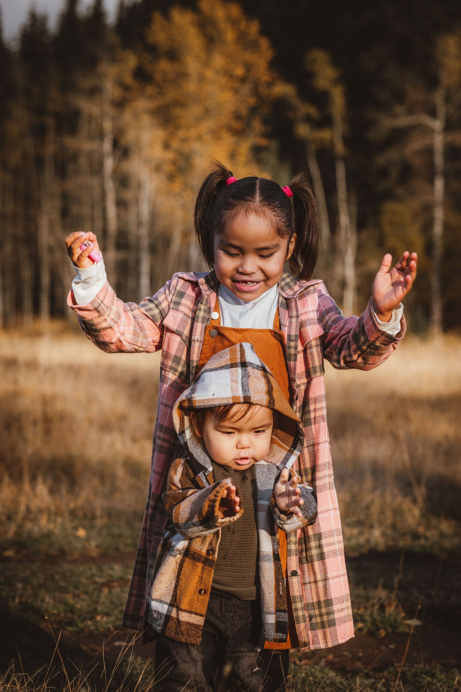 A young girl in a pink plaid shirt laughs while standing behind a toddler in a fall outdoor setting.
