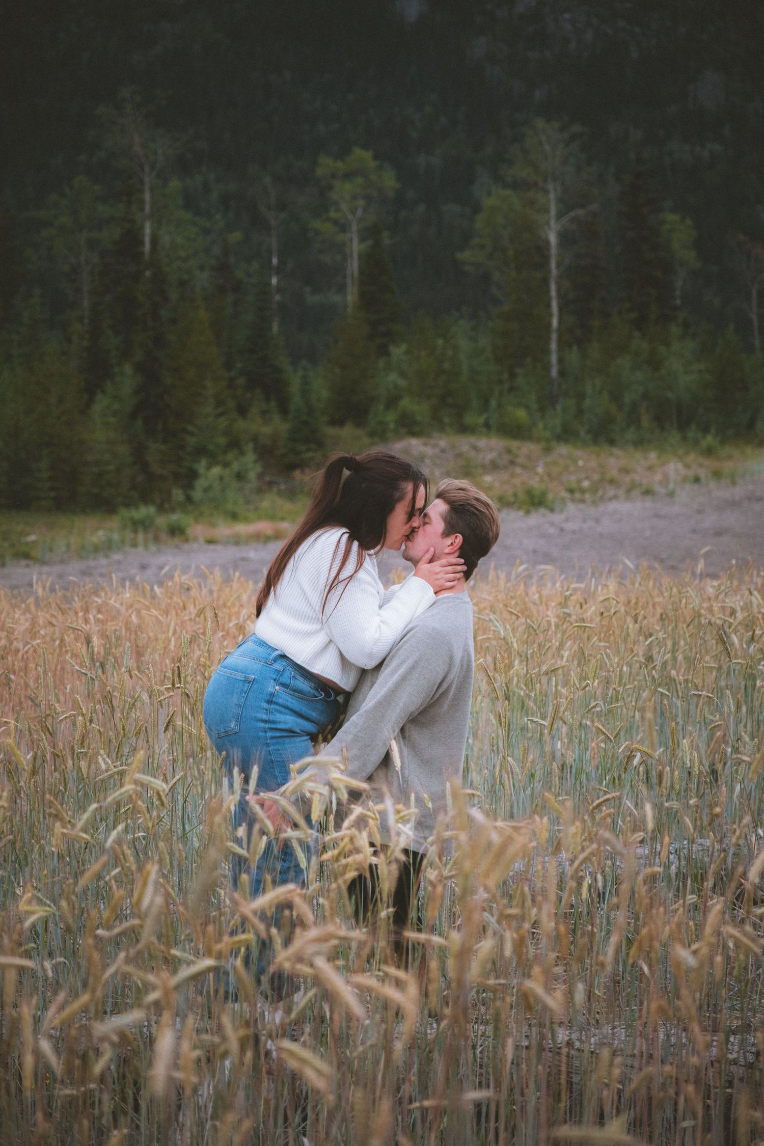 Couple shares a romantic kiss as man lifts woman in a golden wheat field surrounded by forest.