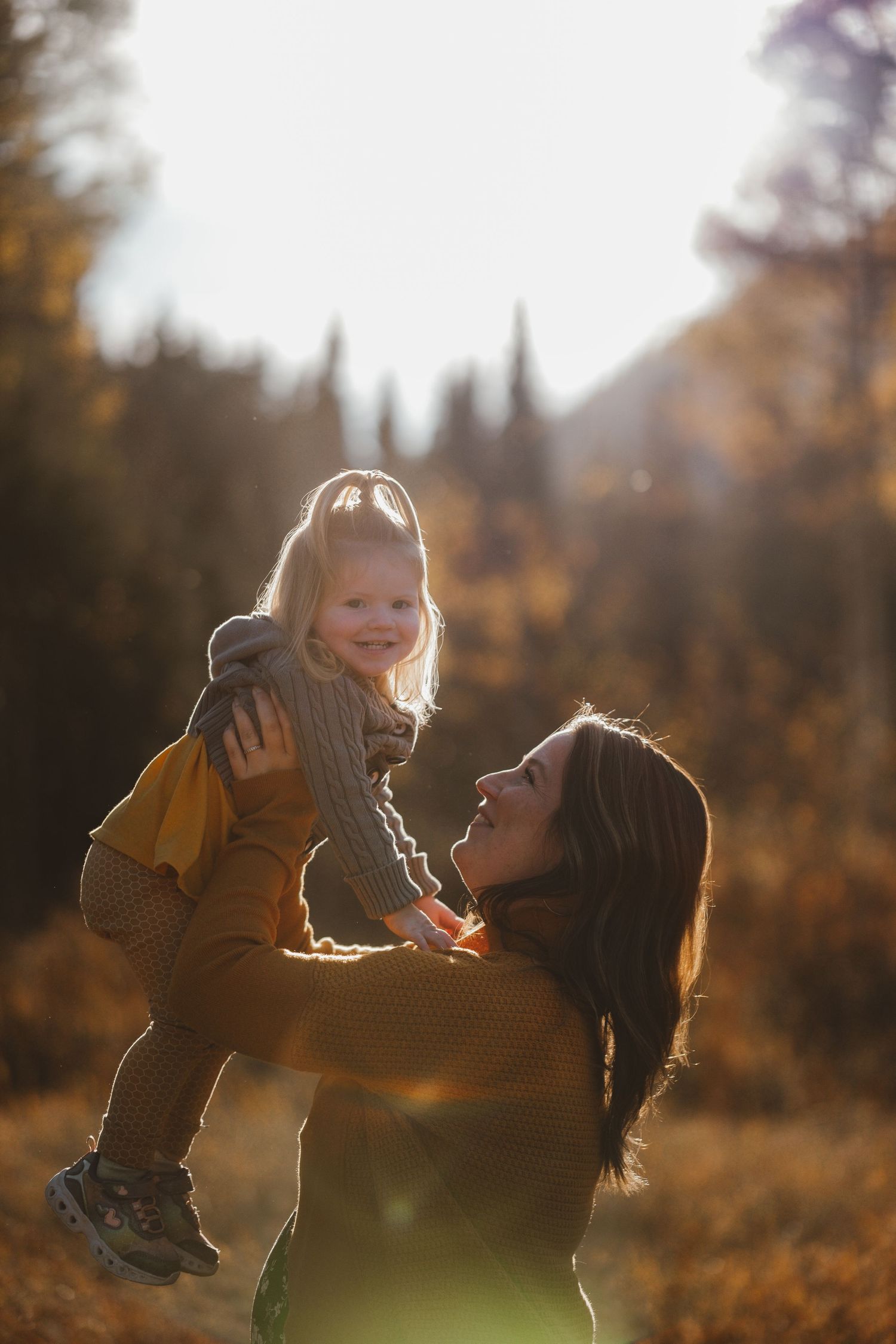 Laughing mother lifting smiling toddler girl in golden autumn sunlight in a meadow.