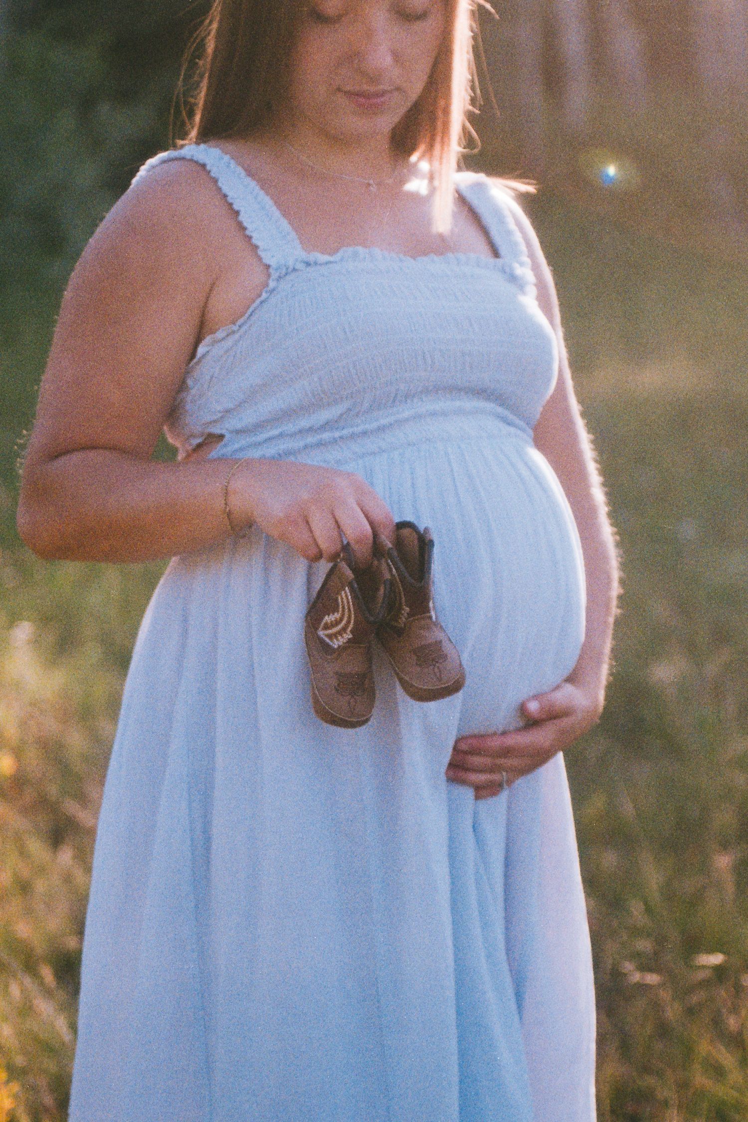 Expectant mother in blue dress holding tiny baby shoes near her pregnant belly outdoors.