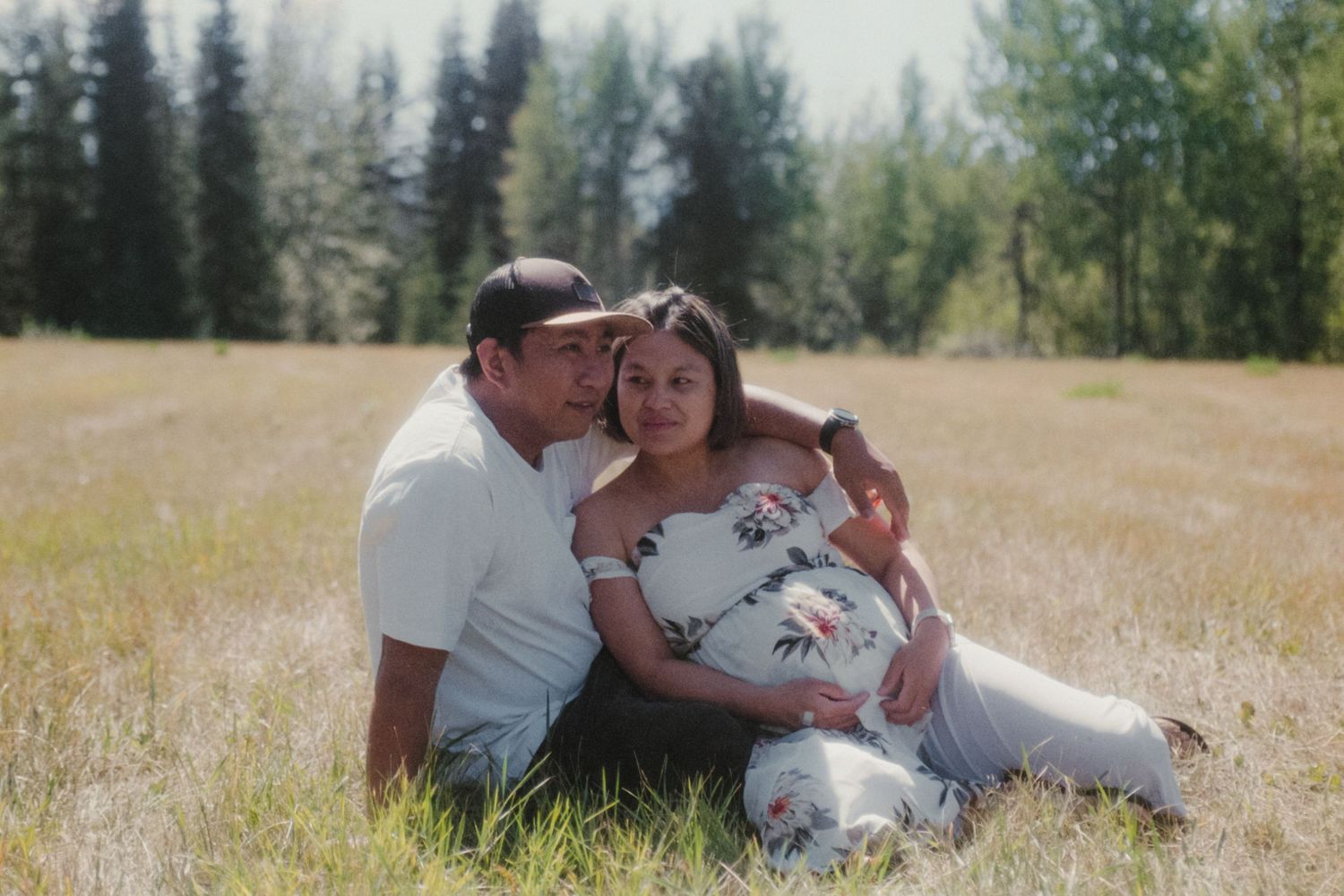 Couple sitting together in a sunny meadow, man embracing pregnant woman in white floral dress.