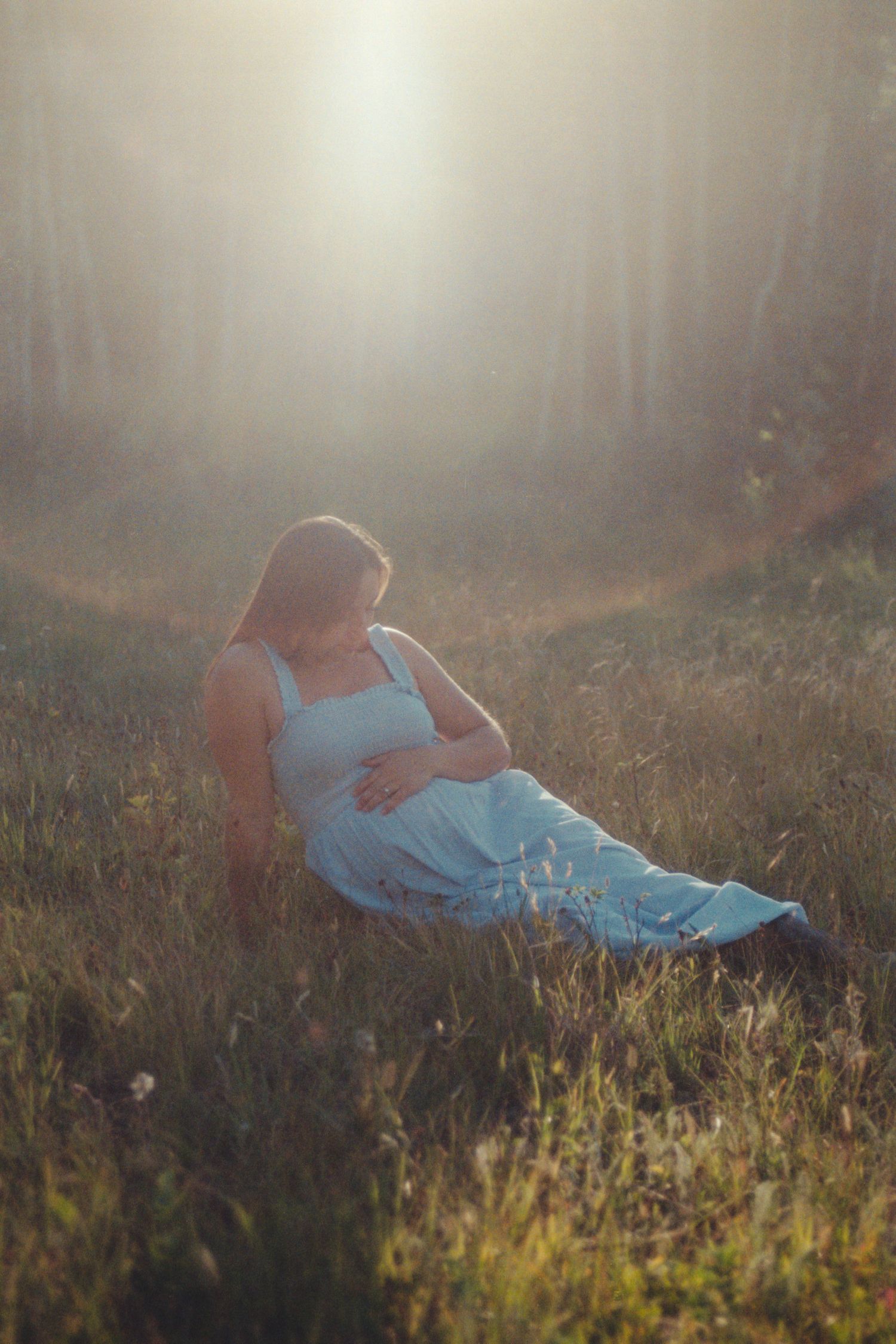 Serene pregnant woman in blue dress sitting in glowing sunlit meadow near birch trees.