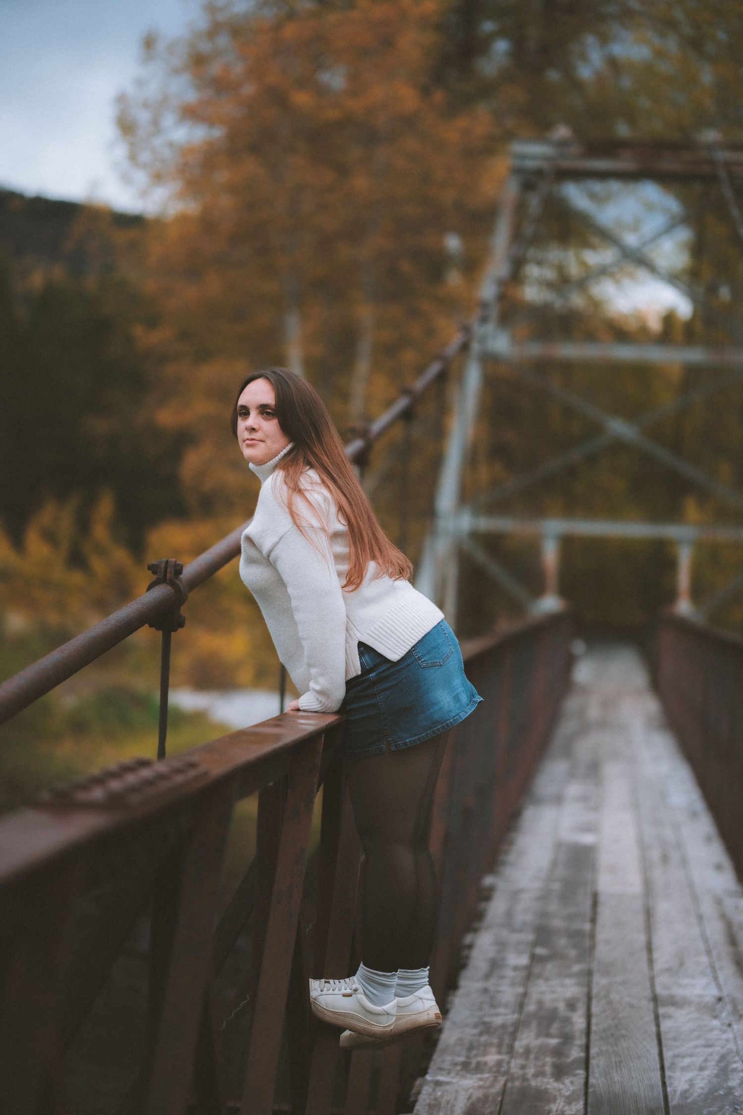 Young woman in white sweater and denim skirt leaning on rustic bridge railing surrounded by autumn foliage.