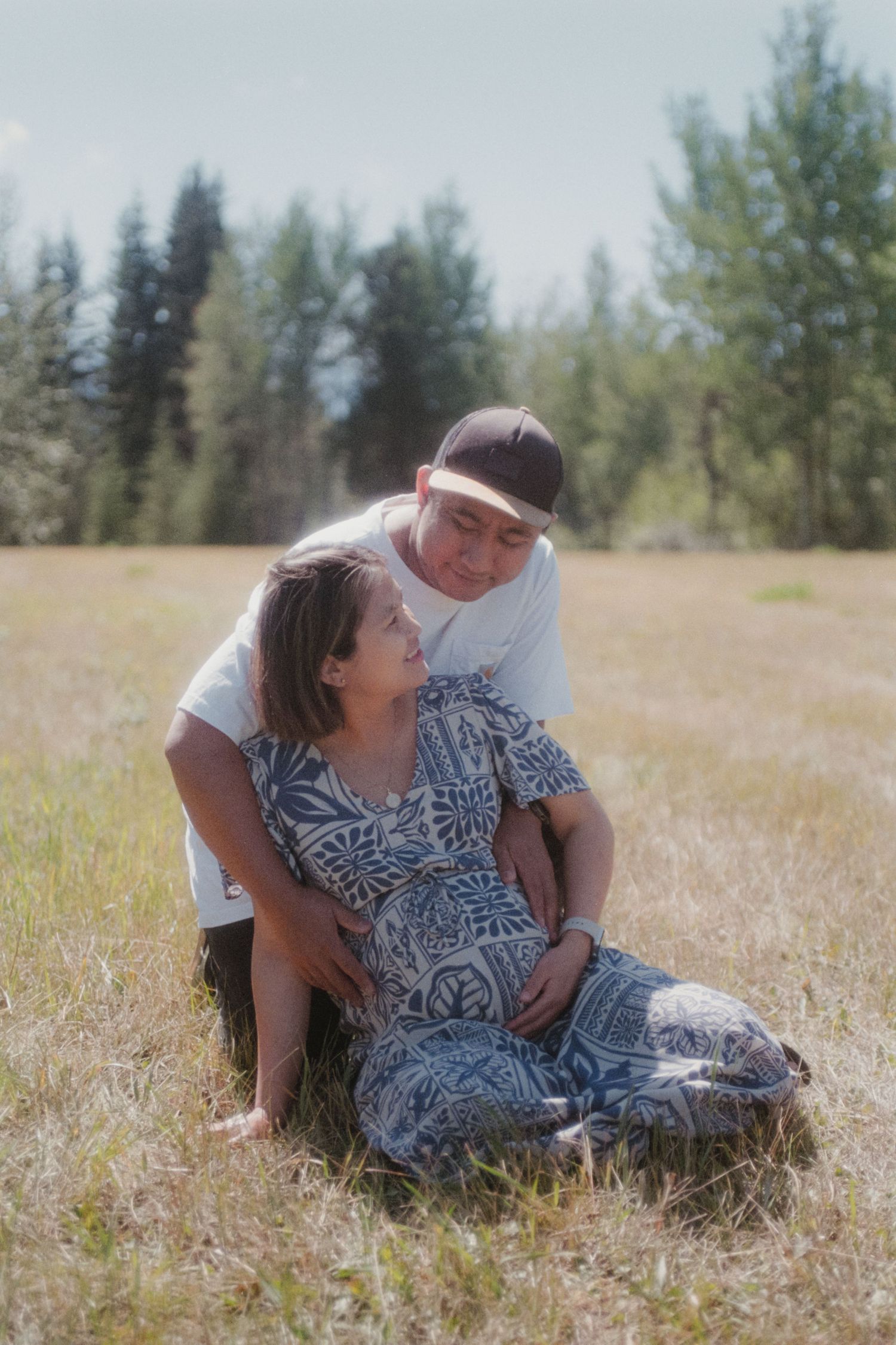 Couple sharing a tender moment in a sunny field, man embracing pregnant woman in blue floral dress.