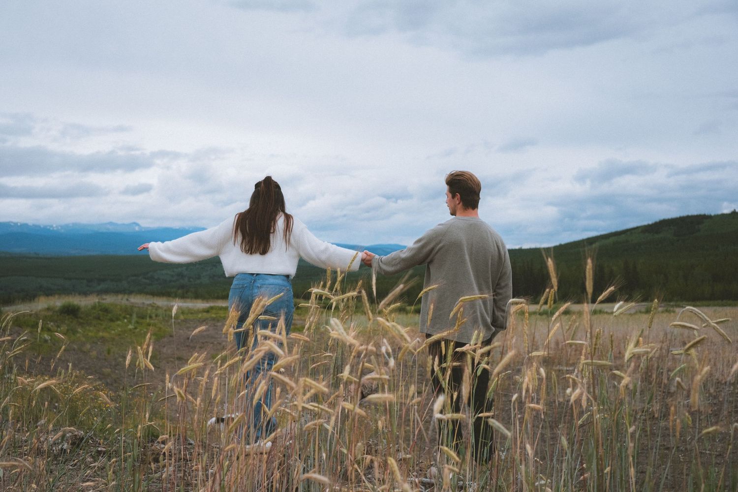 Couple holds hands walking through golden wheat field with mountain views under cloudy sky.