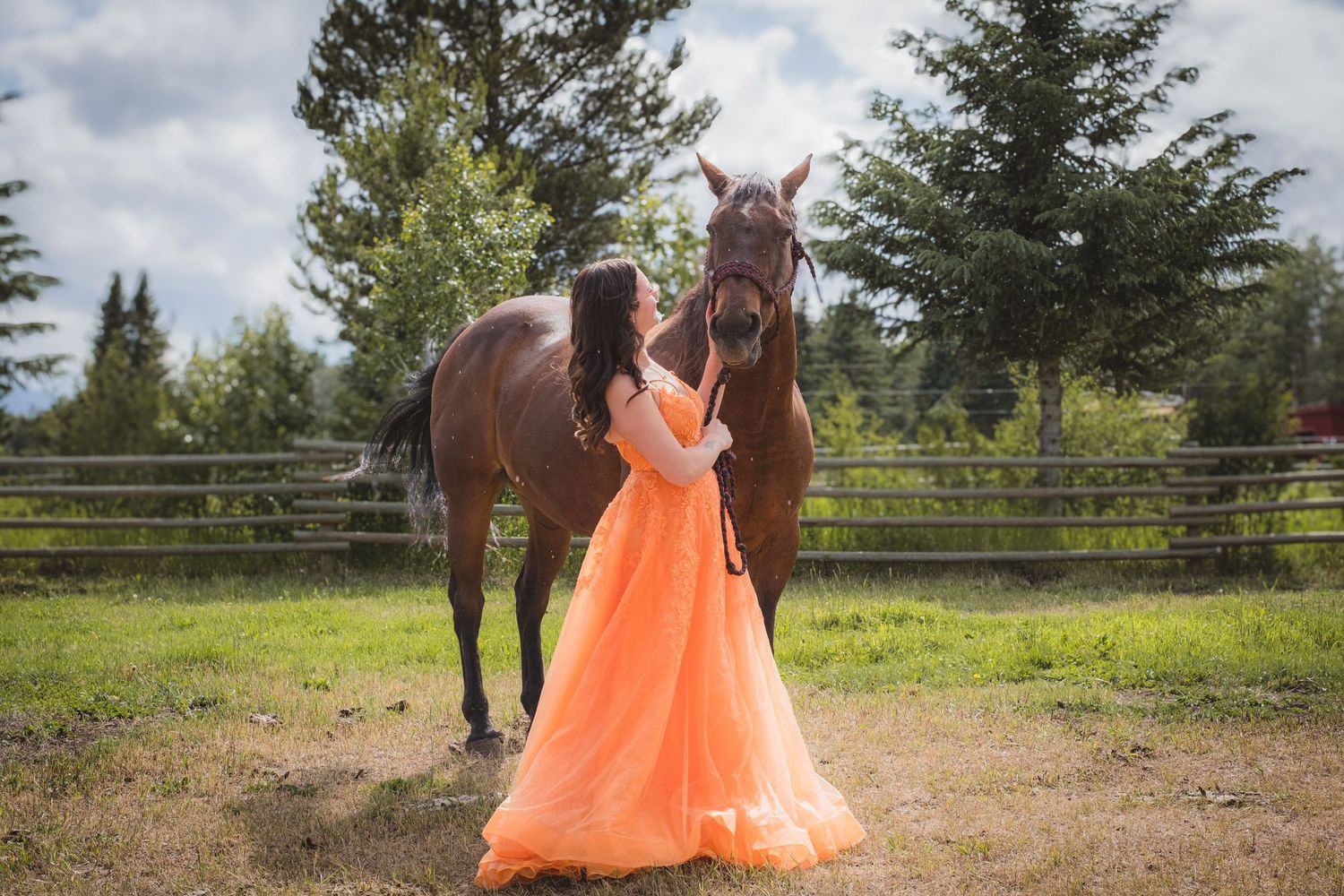 Woman in flowing orange gown embraces dark horse near wooden fence outdoors.