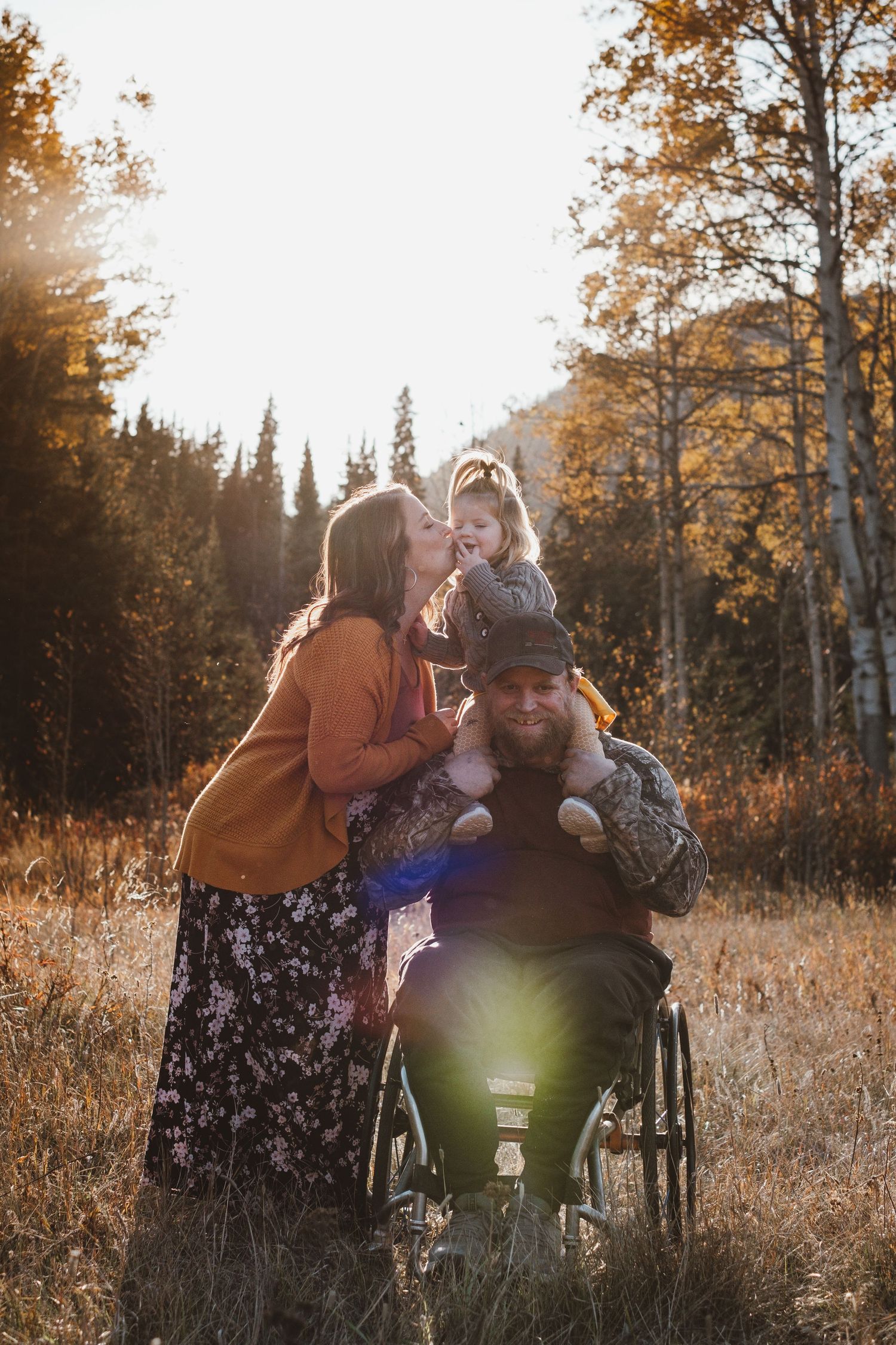 Family portrait with mother kissing toddler and father in wheelchair among golden autumn aspens.