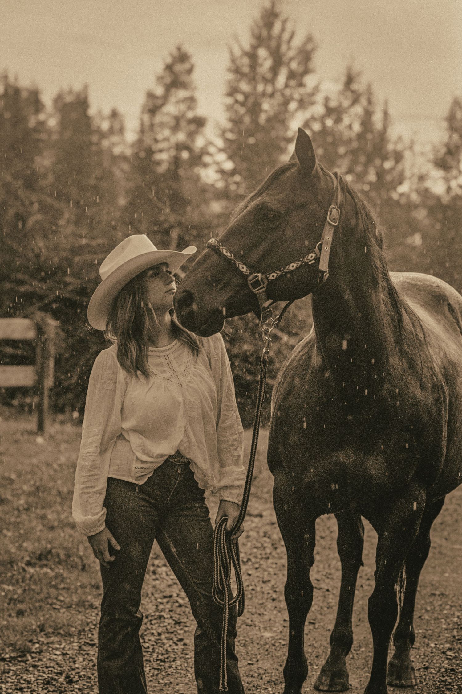 Sepia-toned photo of a woman in a cowboy hat and jeans standing with a dark horse in a rainy outdoor setting.