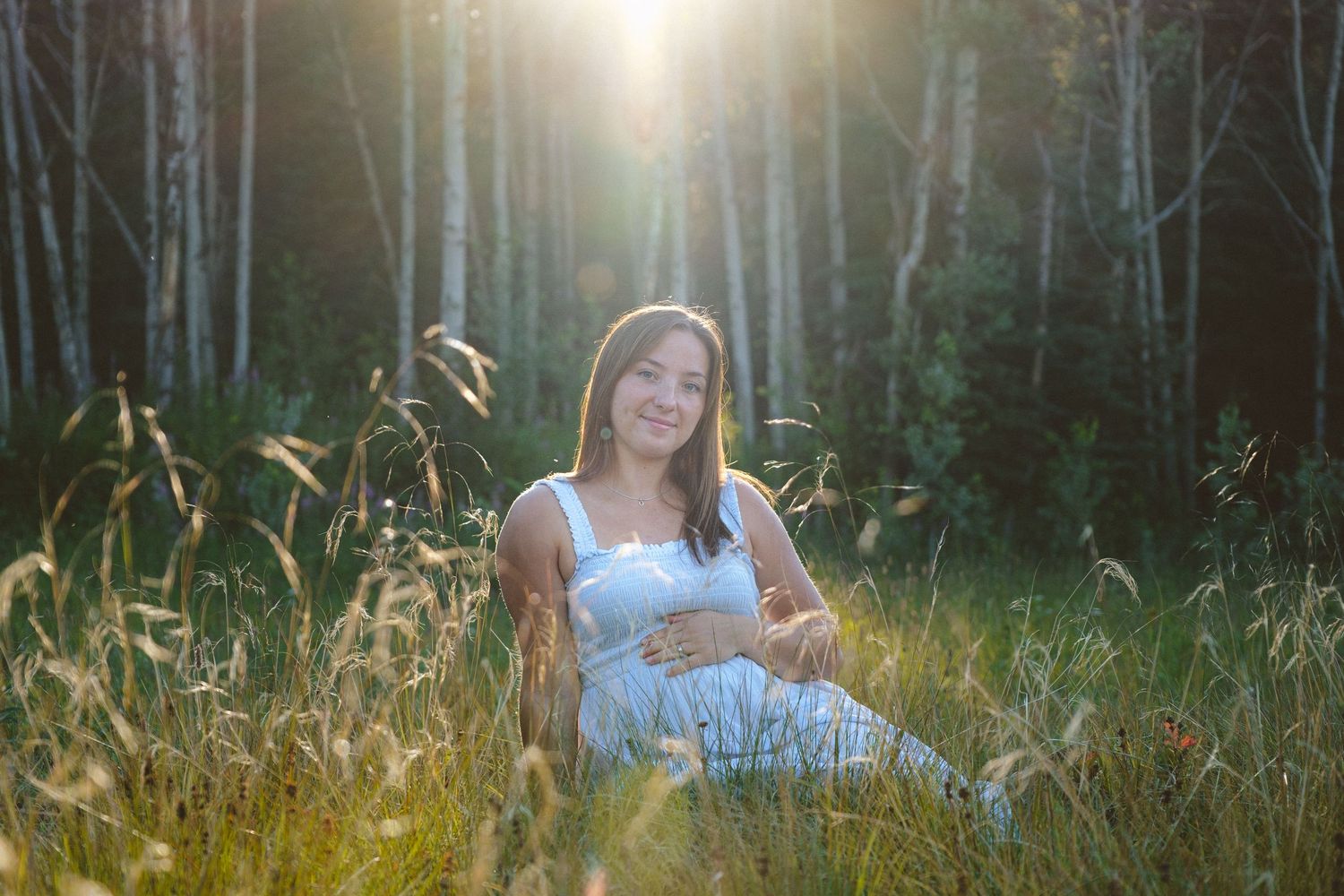 Pregnant woman in white dress sitting in tall grass with warm sunlight glowing through trees.