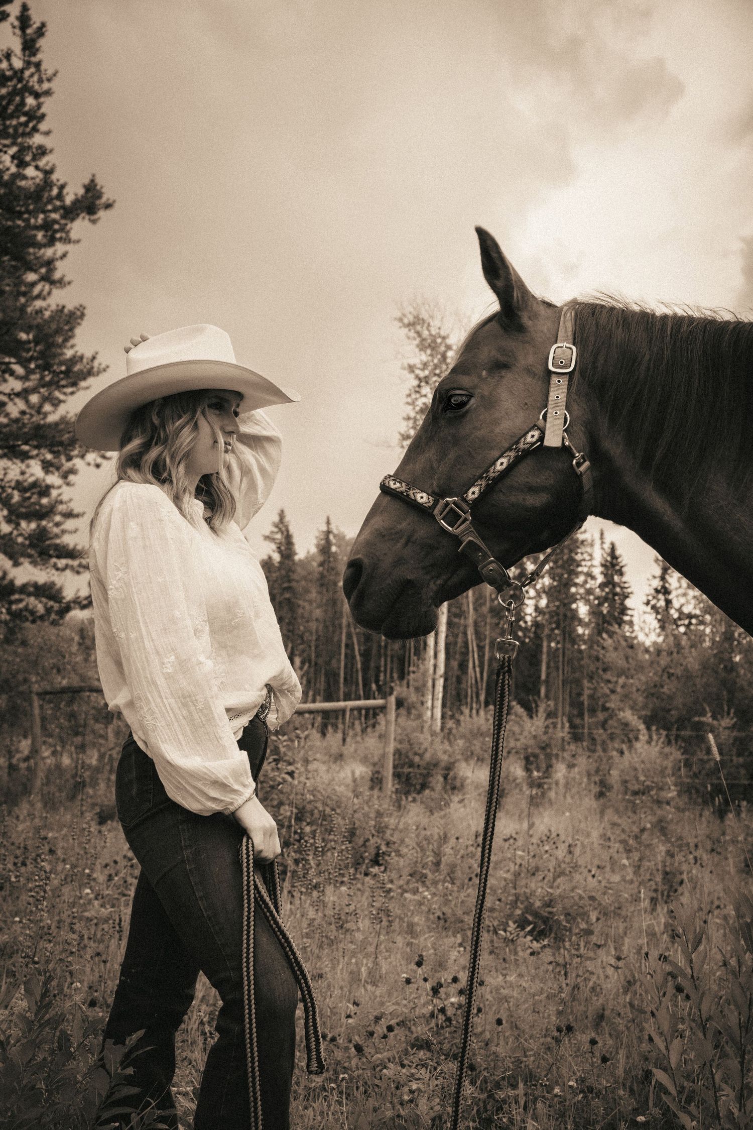 Sepia-toned photo of a blonde woman in a cowboy hat and white blouse standing face-to-face with a dark horse in a field.