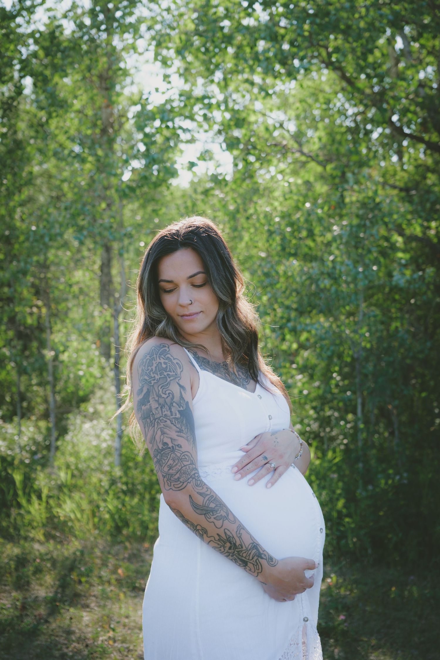 Tattooed pregnant woman in white dress gently holding baby bump surrounded by lush green trees.