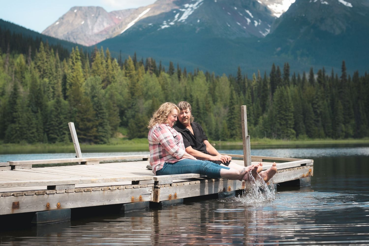 Two women sitting on lakeside dock splashing feet in water with forested mountains in background.