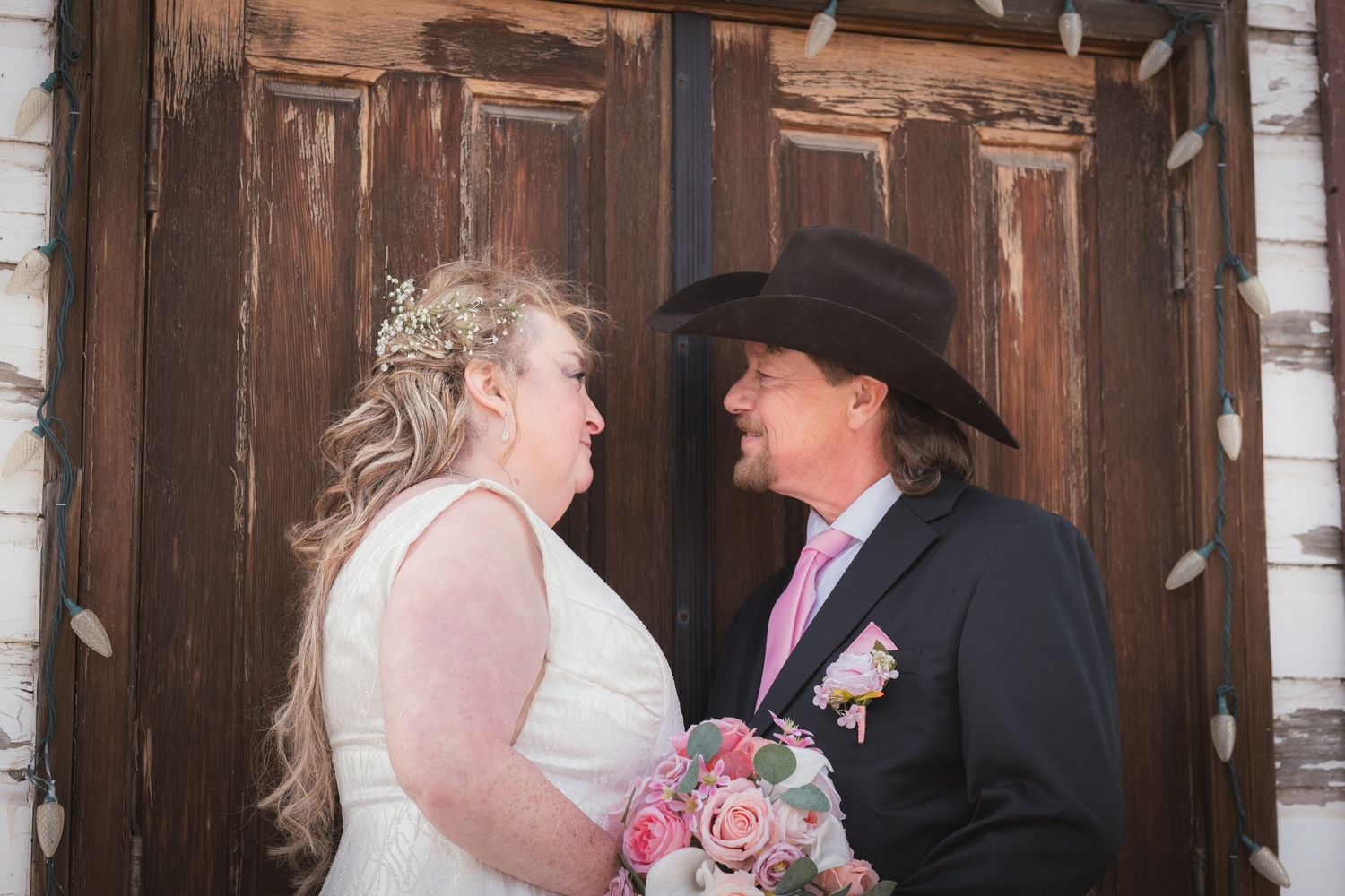 Bride and groom in cowboy hat share a loving gaze before rustic wooden doors on their wedding day.