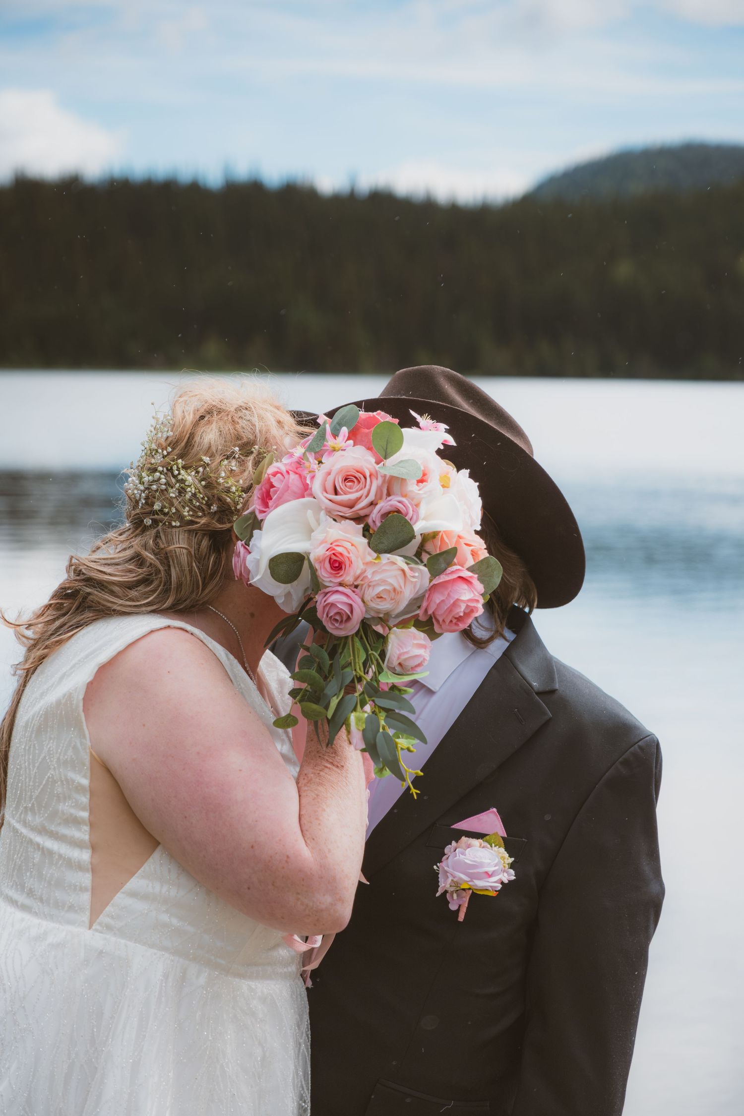 Bride and groom kissing behind pink rose bouquet at lakeside wedding.