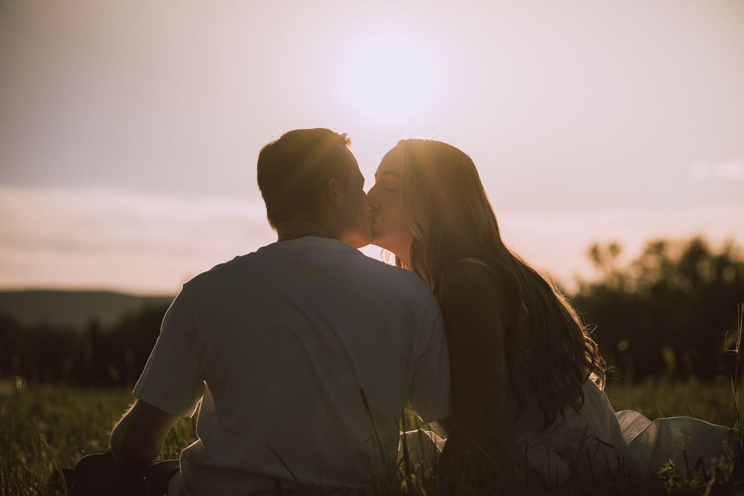 Couple sharing a tender kiss silhouetted against a golden sunset in an open grassy field.