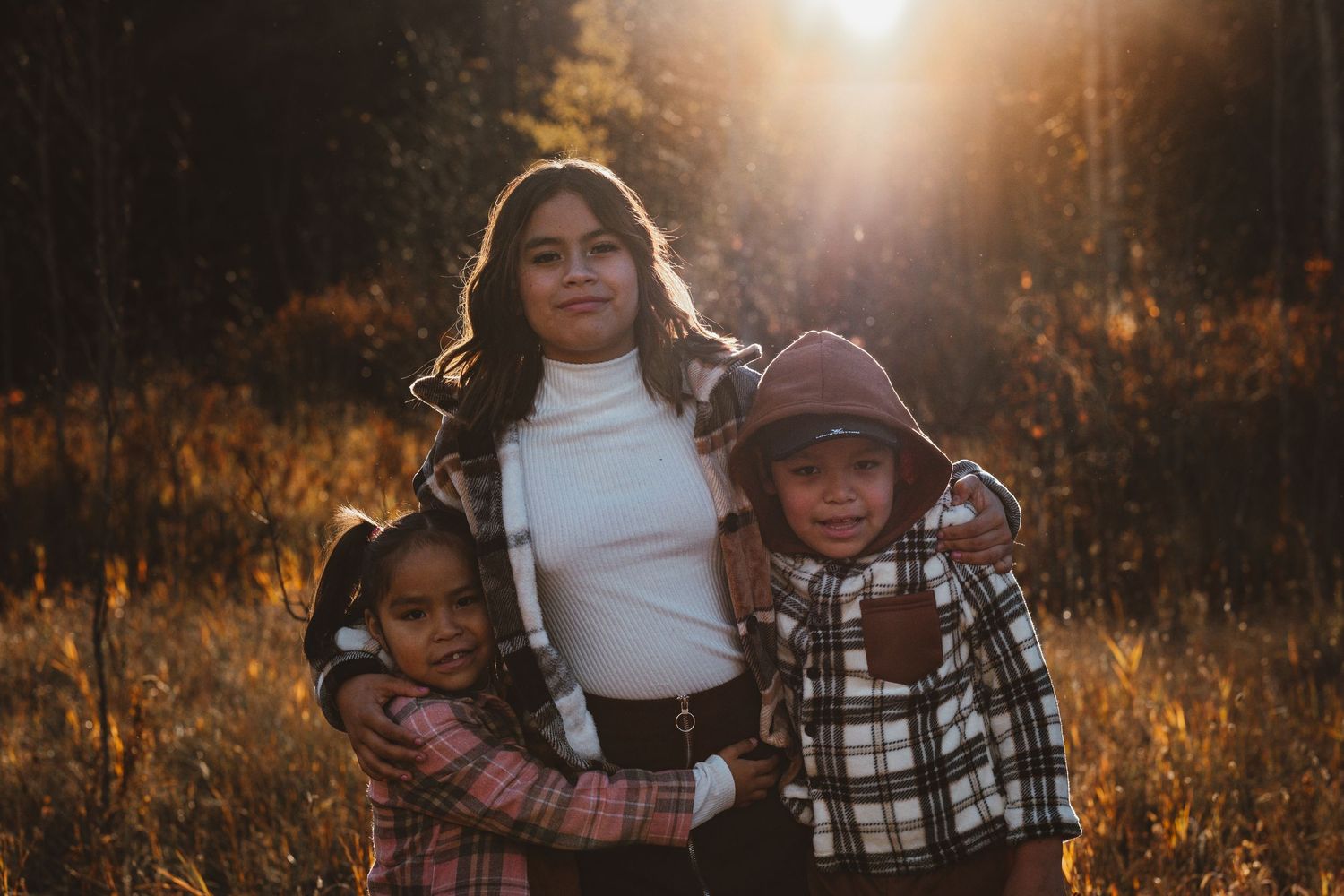 A teenage girl hugs two younger siblings in a warm golden autumn forest setting.