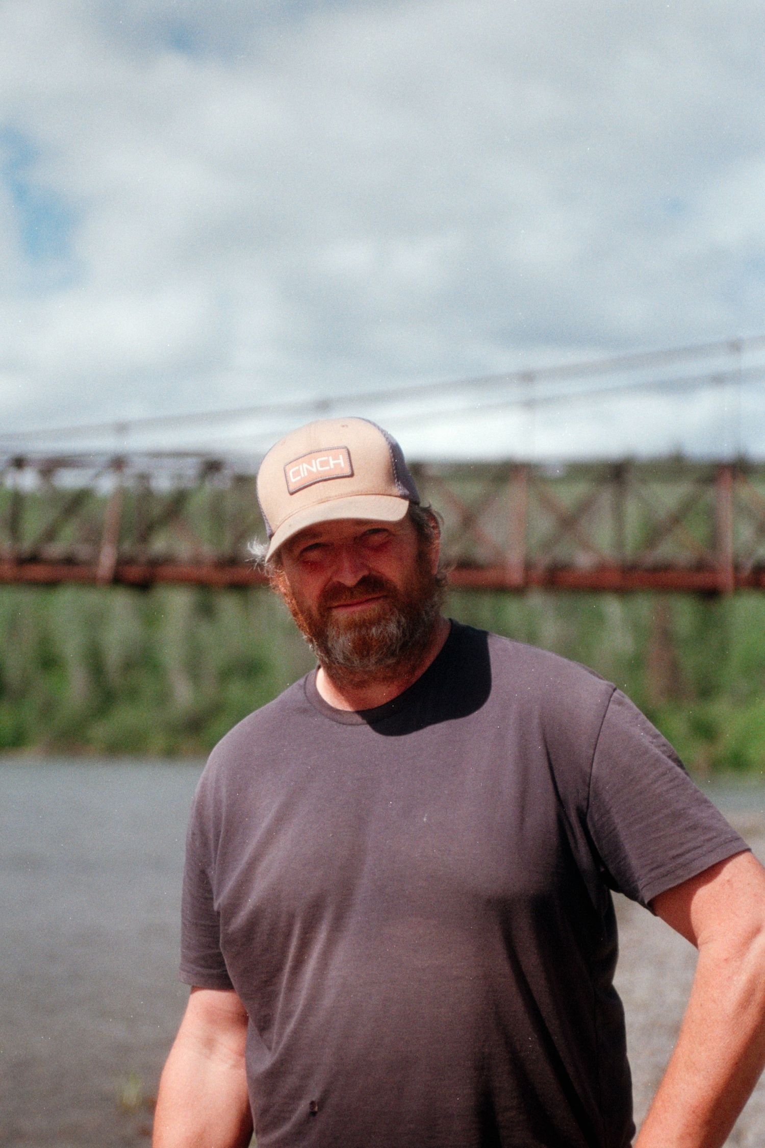 Bearded man in gray t-shirt and cap standing near a river with an old iron bridge behind him.