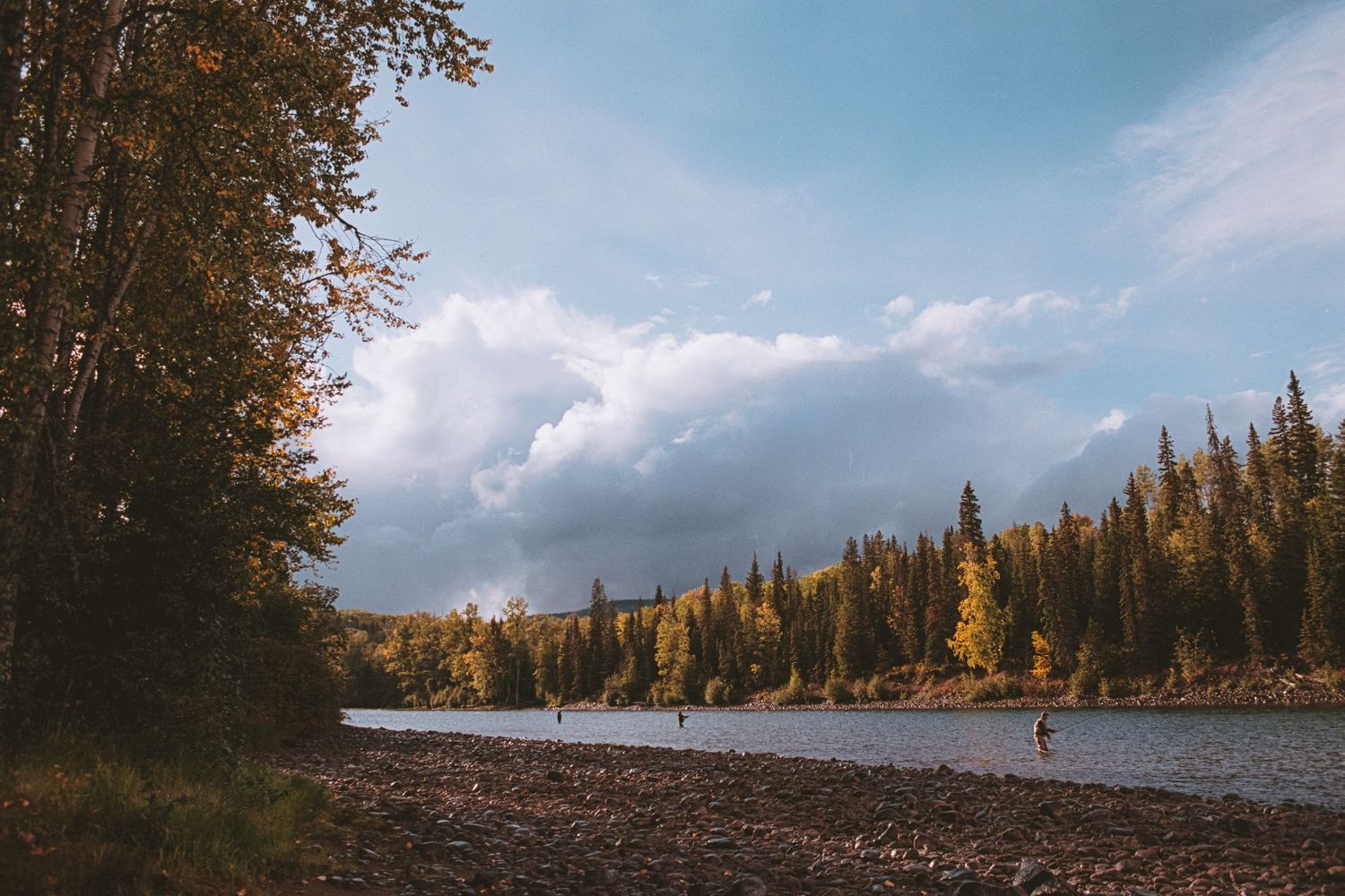 People fishing along a rocky riverbank surrounded by lush evergreen forest under dramatic cloudy skies.