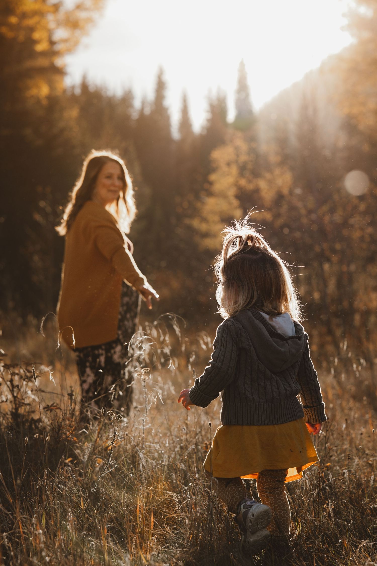 Young girl in yellow skirt walking toward mother through golden autumn meadow grass.