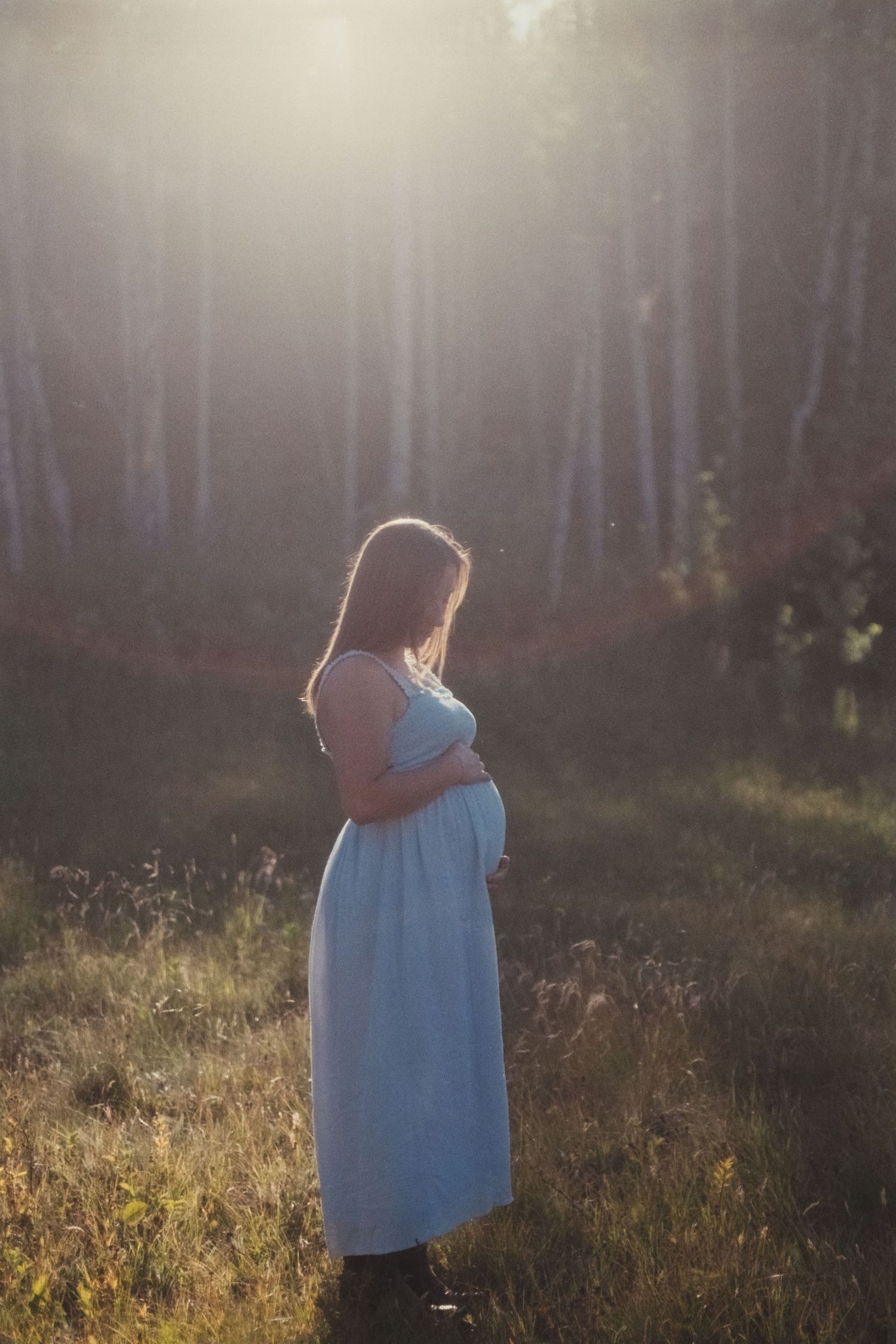 Side profile of pregnant woman in blue dress gently holding bump in radiant forest light.