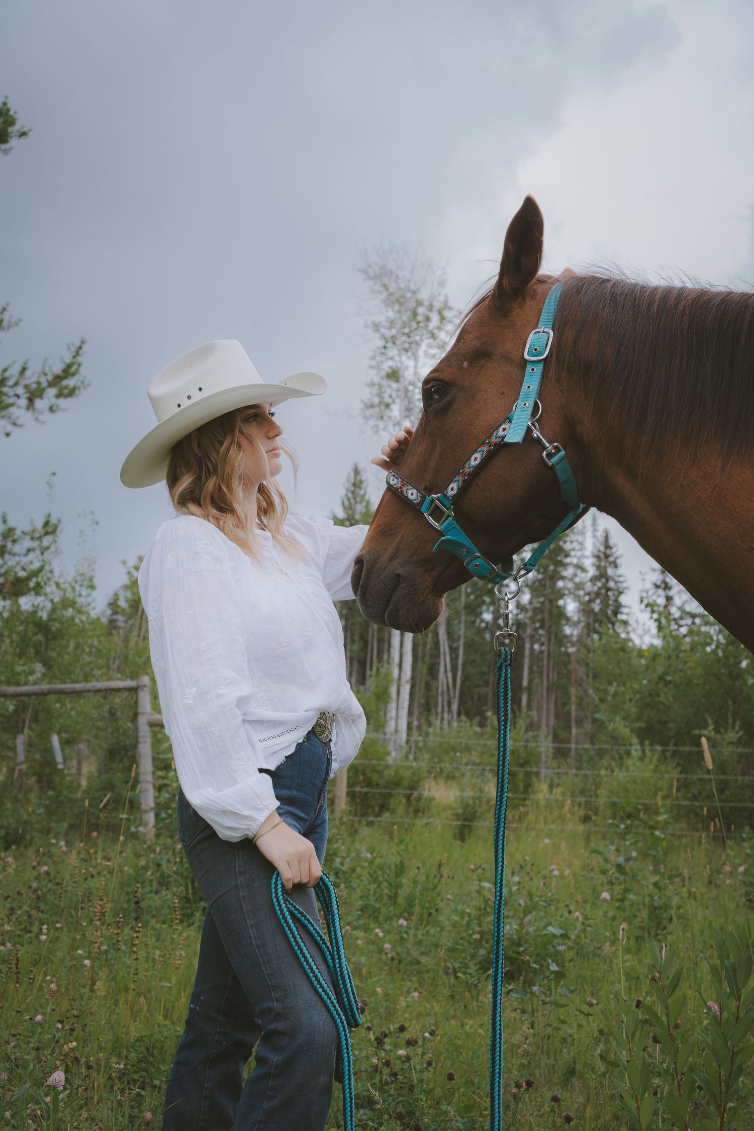 Woman in white blouse and cowboy hat stands face-to-face with a chestnut horse wearing a teal halter outdoors.