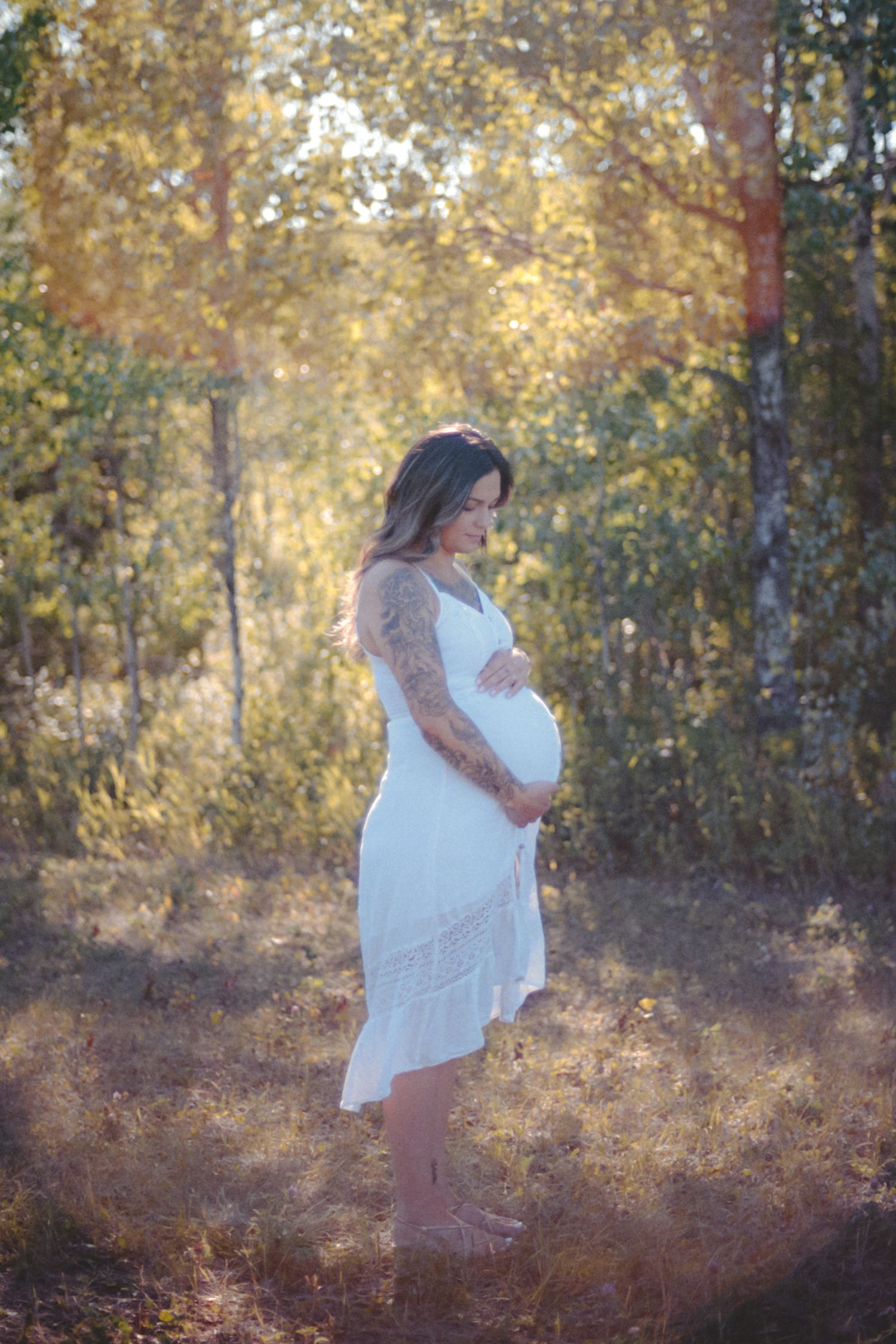 Tattooed pregnant woman in white lace dress standing in sunlit autumn forest, holding baby bump.