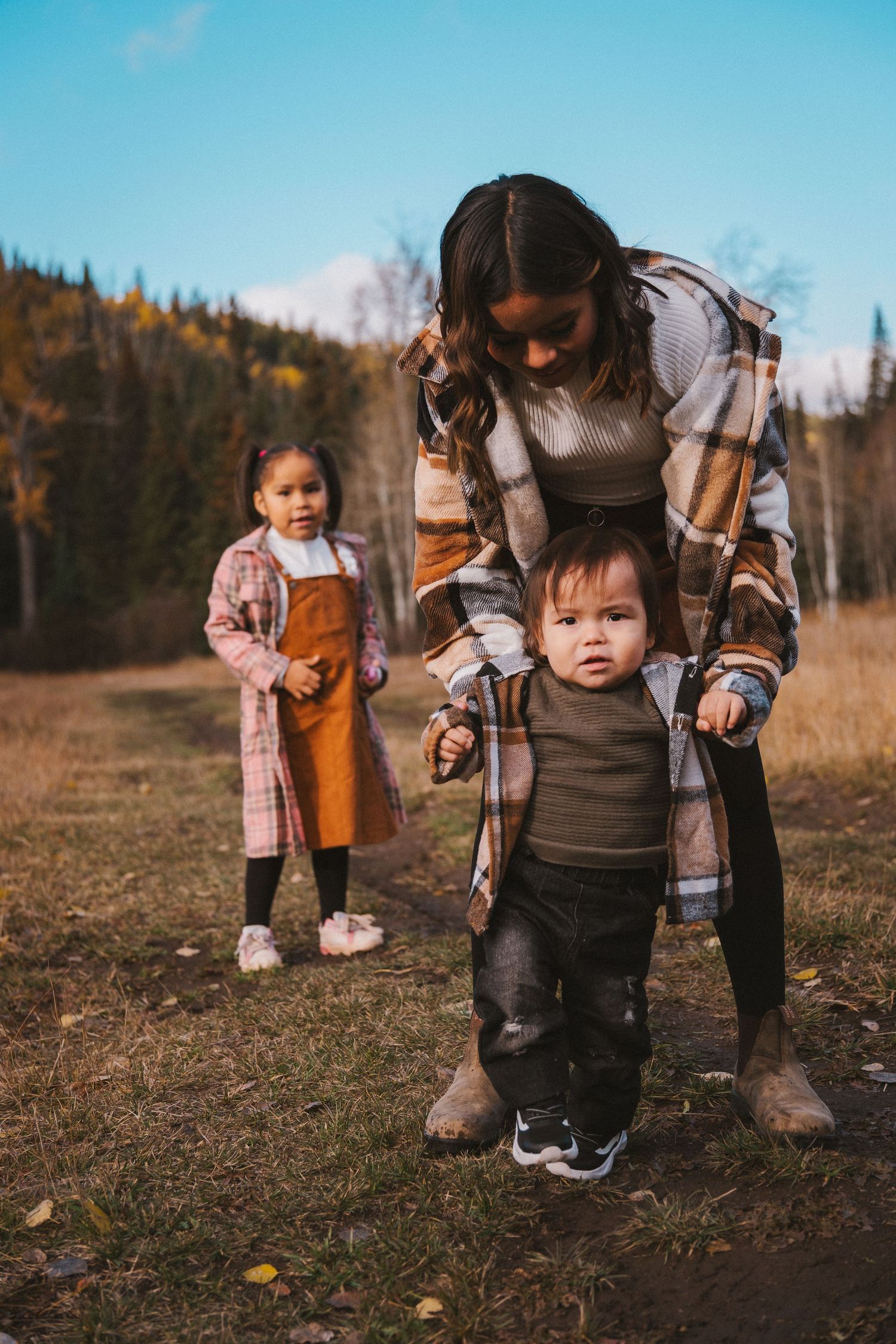 Mother helping baby take first steps outdoors in autumn while toddler girl watches nearby.