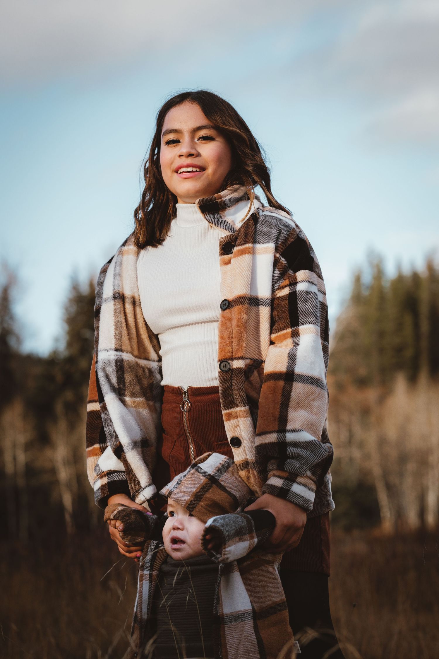 A teenage girl in a brown plaid coat and white turtleneck smiles in an autumn field.