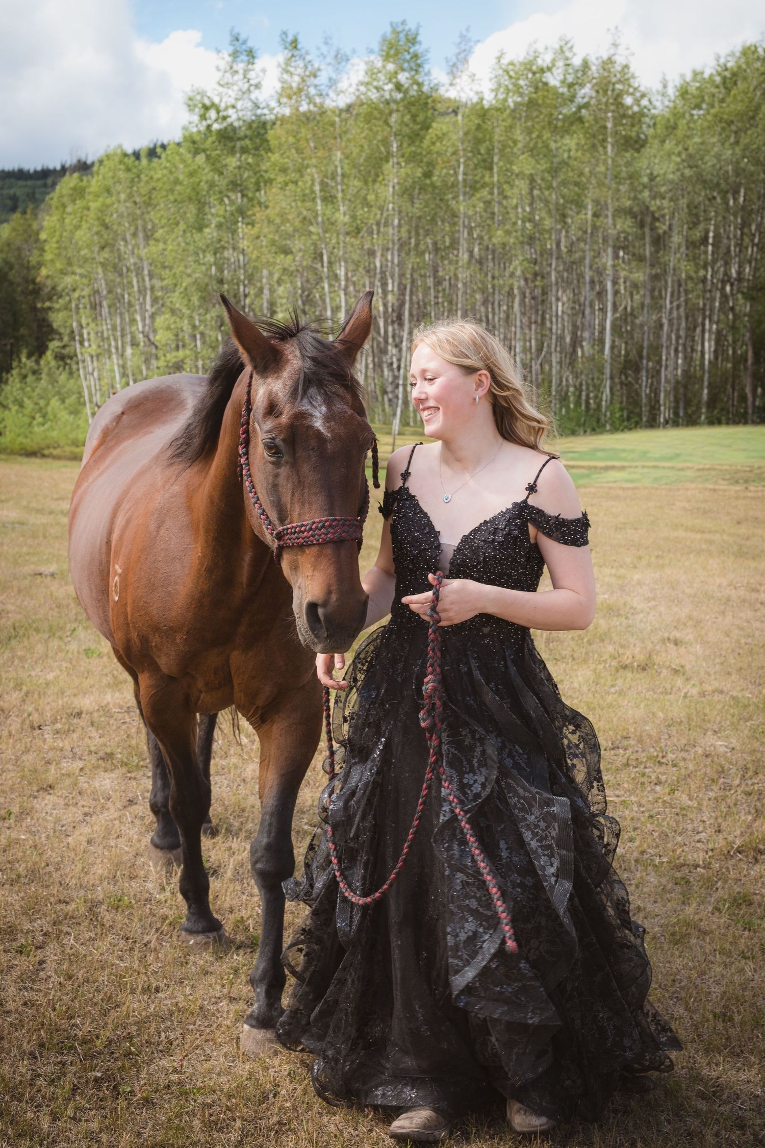 Young blonde woman in black lace gown walking with chestnut horse in aspen tree meadow.