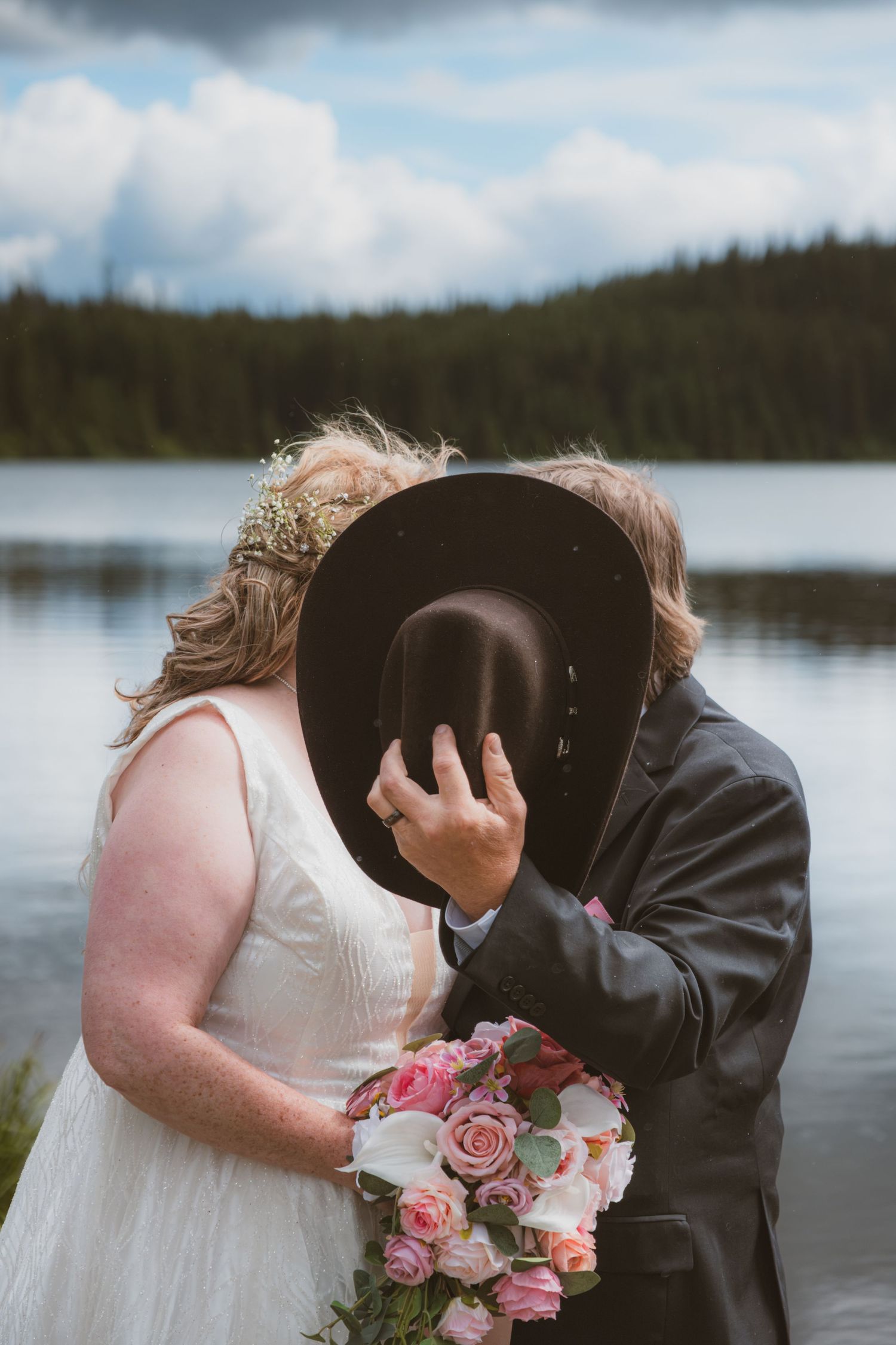 Couple hiding kiss behind cowboy hat at lakeside wedding photo session.