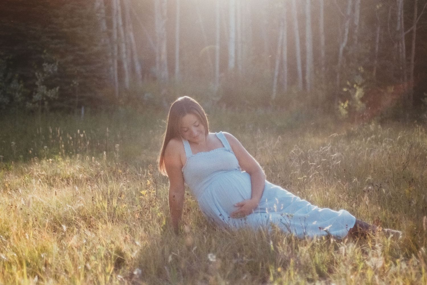 Pregnant woman in white dress sitting in sunlit field near birch trees.