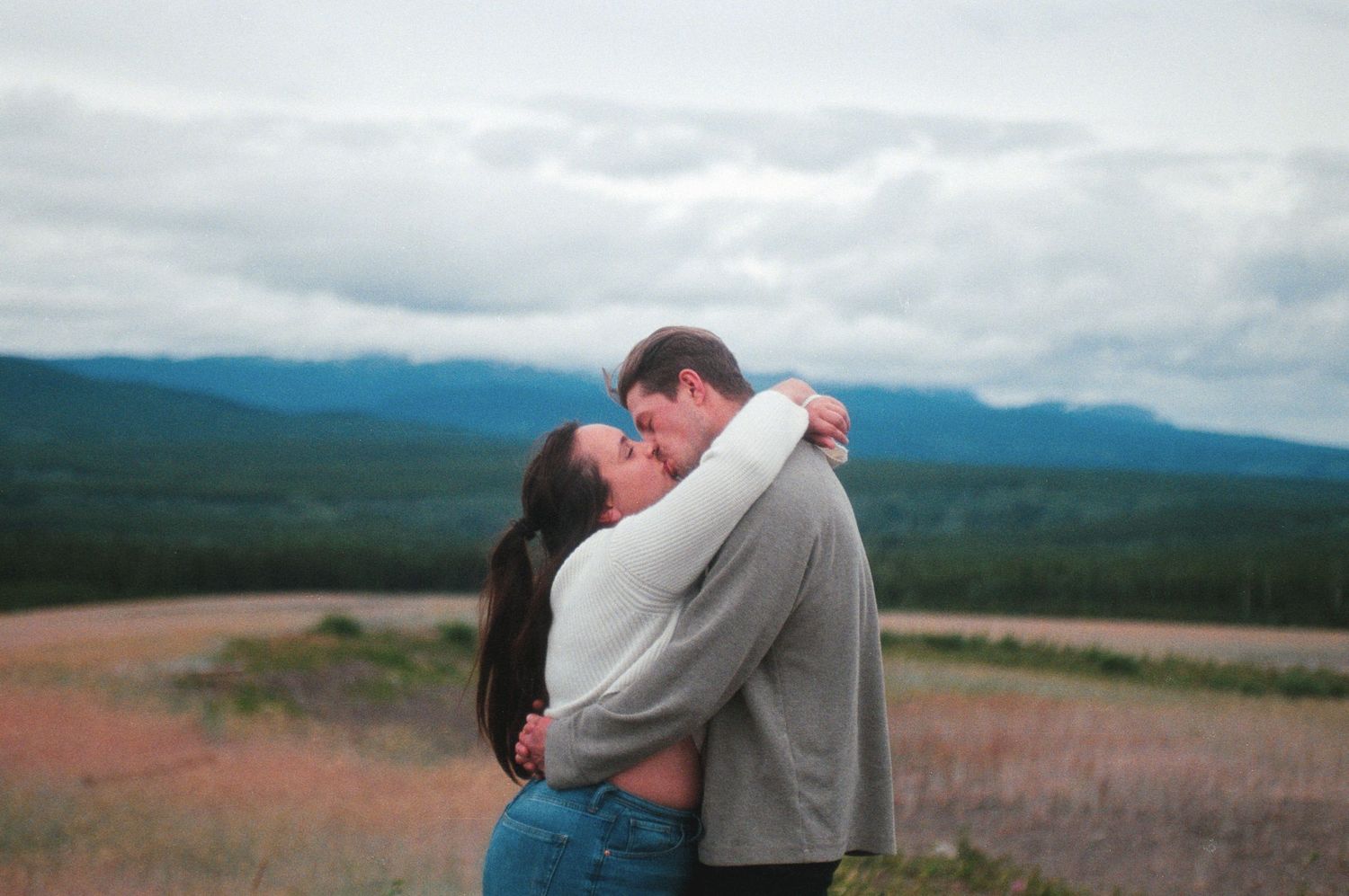 Couple kissing and embracing in an open field with stunning mountain range in the background.