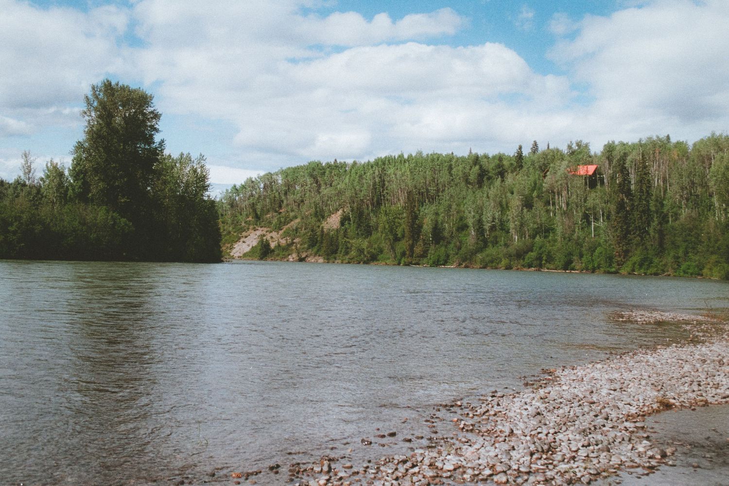 A peaceful river scene with a rocky shoreline, dense forest, and a small red cabin visible on the hill.