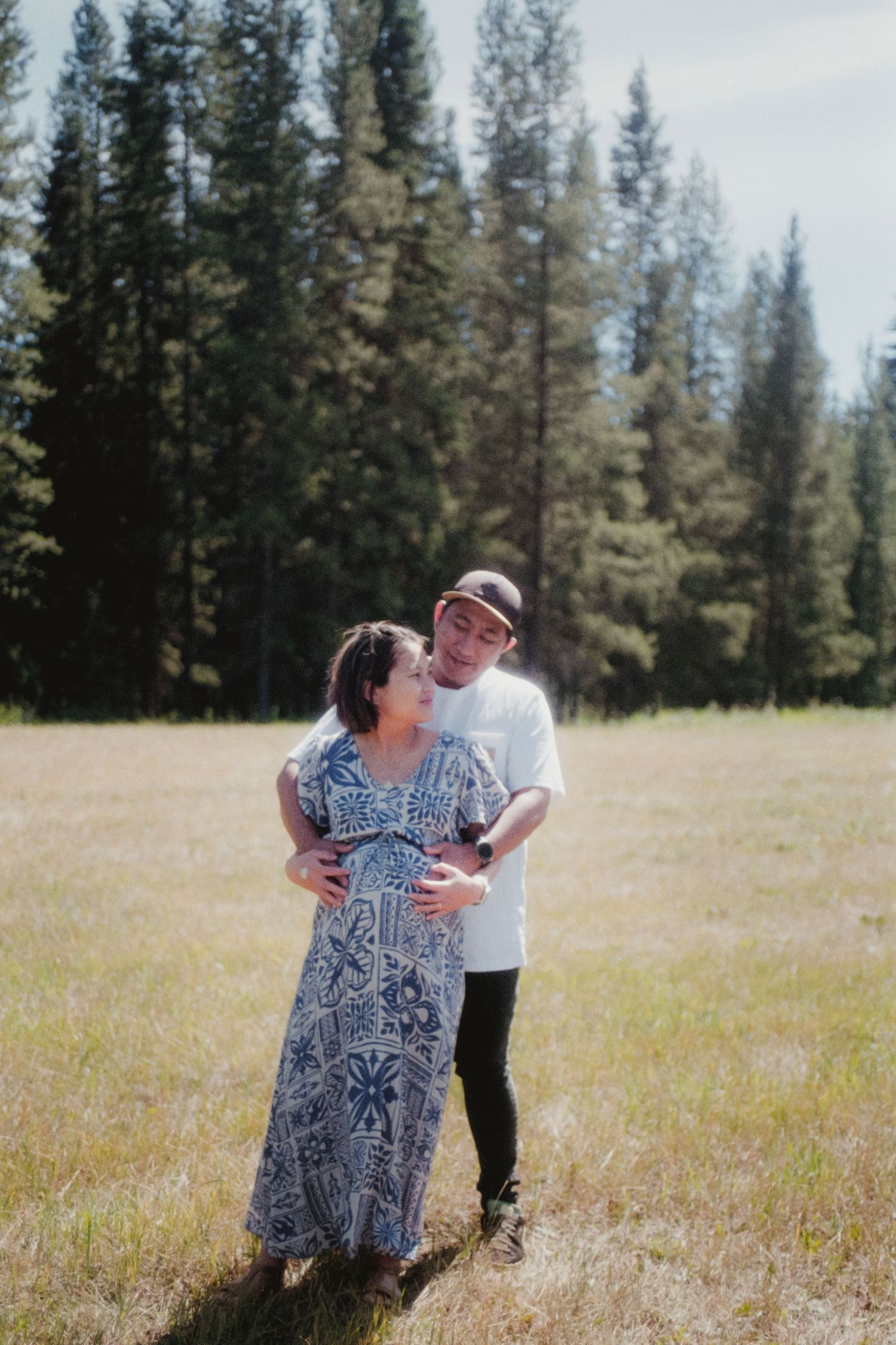 Couple embracing in grassy meadow surrounded by tall pine trees during maternity photoshoot.